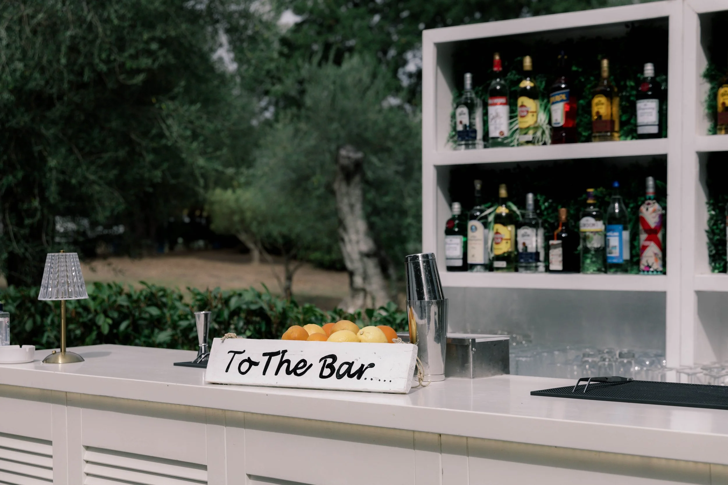 An outdoor bar setup with a white counter, a small lamp, a sign saying 'To The Bar,' a bowl of oranges and lemons, and a shelf filled with various bottles of alcohol, surrounded by greenery.