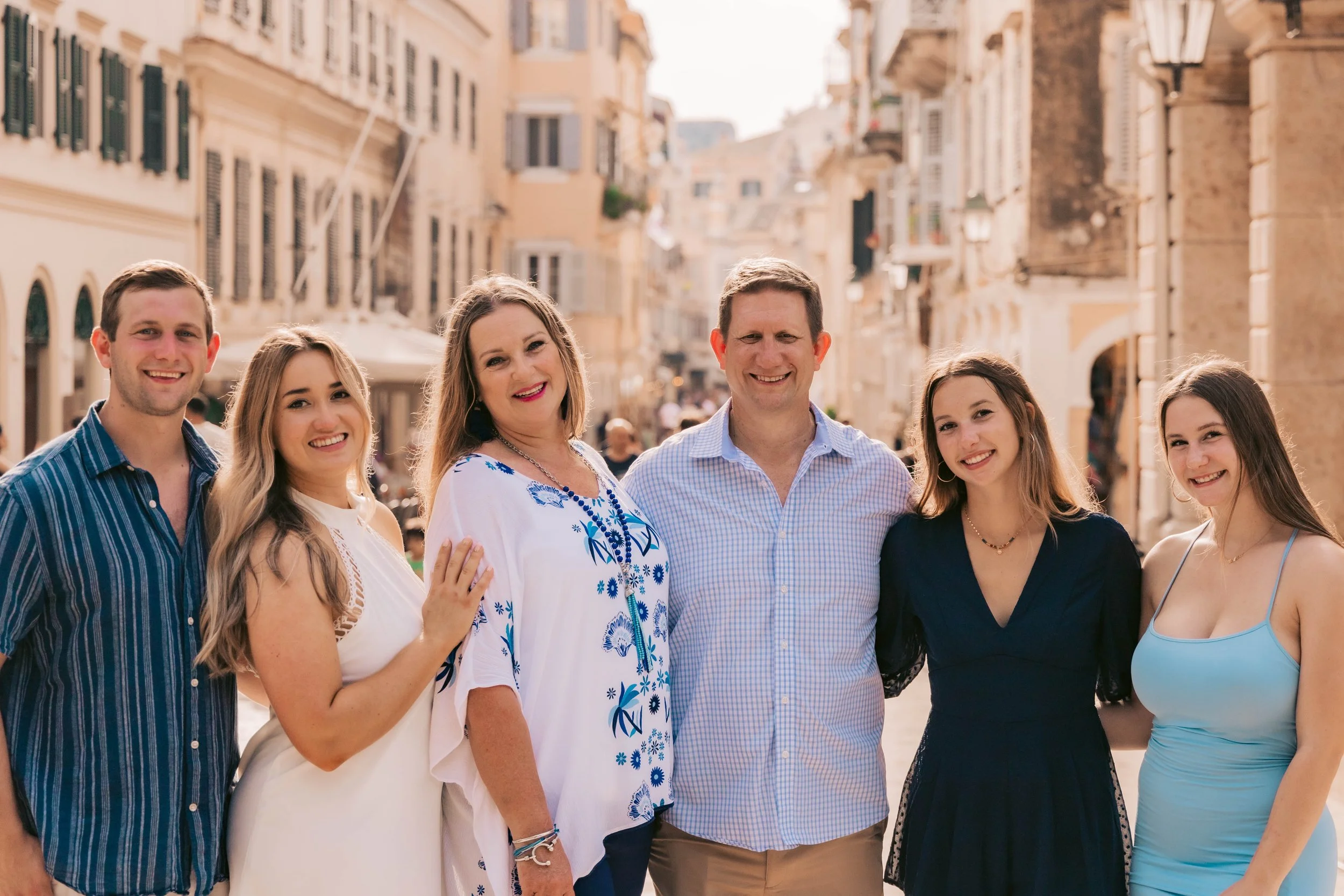 Family of six standing together on a sunny street in a European city, smiling and posing for a photo.