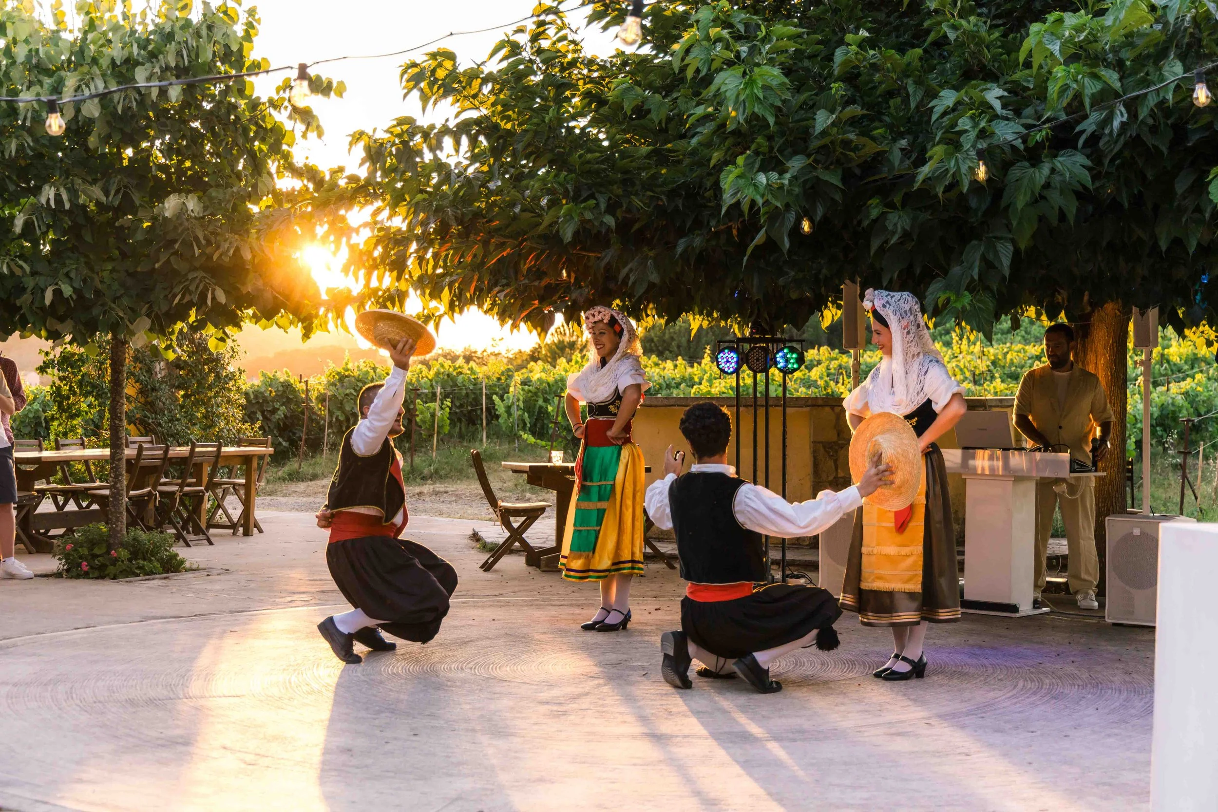 People dressed in traditional folk costumes performing a dance outdoors at sunset, with a DJ in the background and string lights overhead.