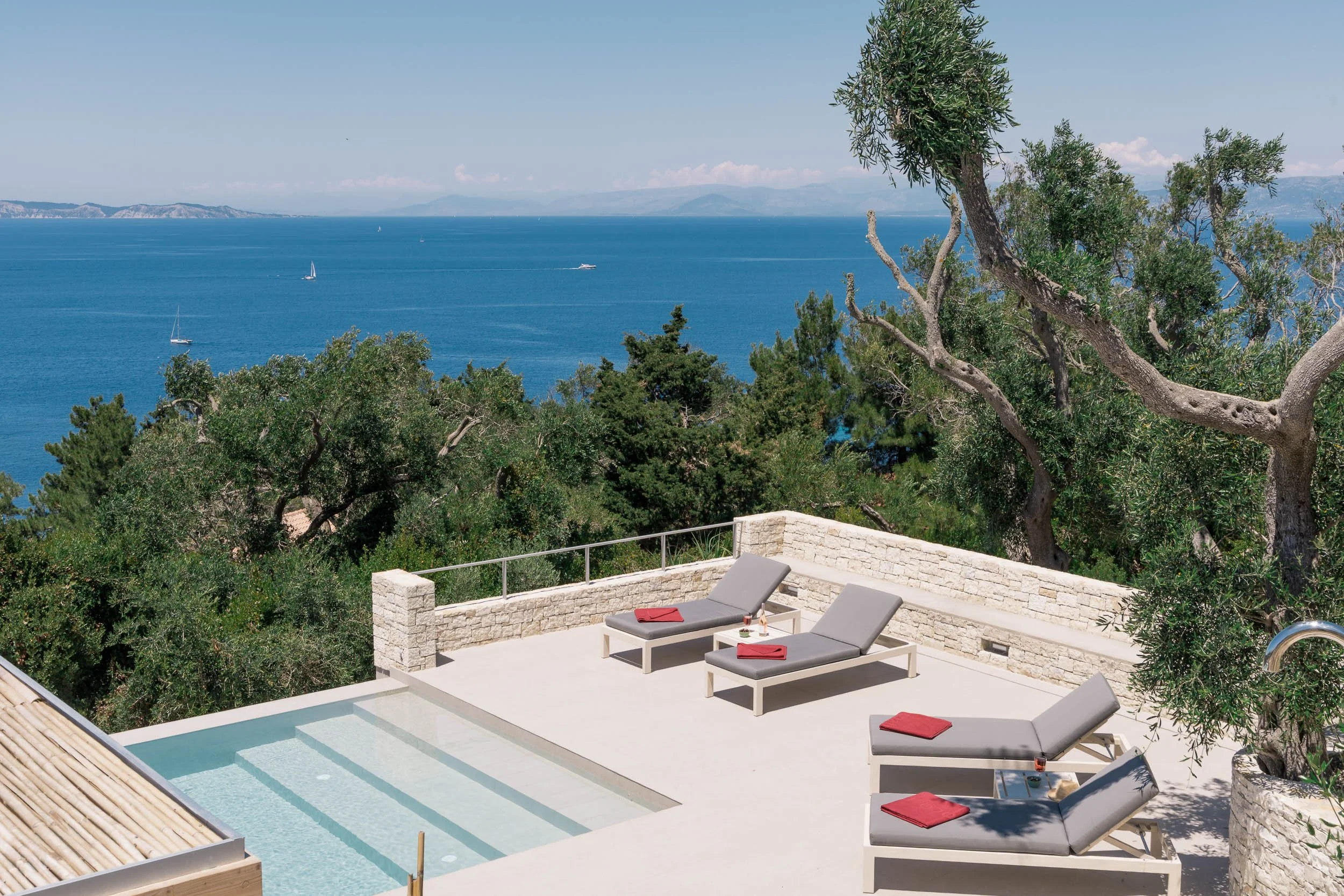 A terrace with three lounge chairs with red towels, a small pool, and a view of the ocean with sailboats and mountains in the distance.