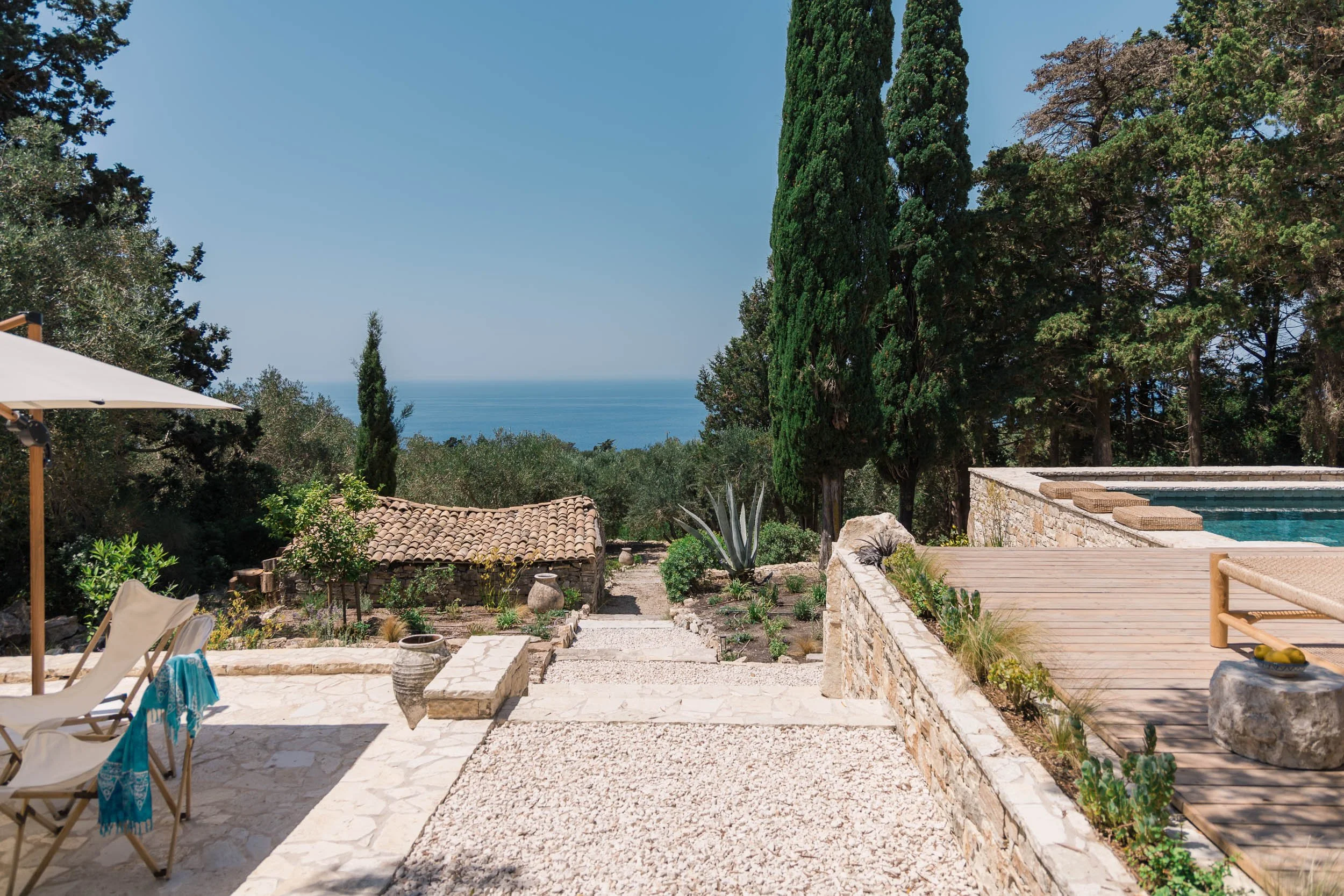 An outdoor patio area with a gravel walkway, surrounded by lush green trees, with a view of the ocean in the background. There are chairs, an umbrella, a small pool, and various potted plants.