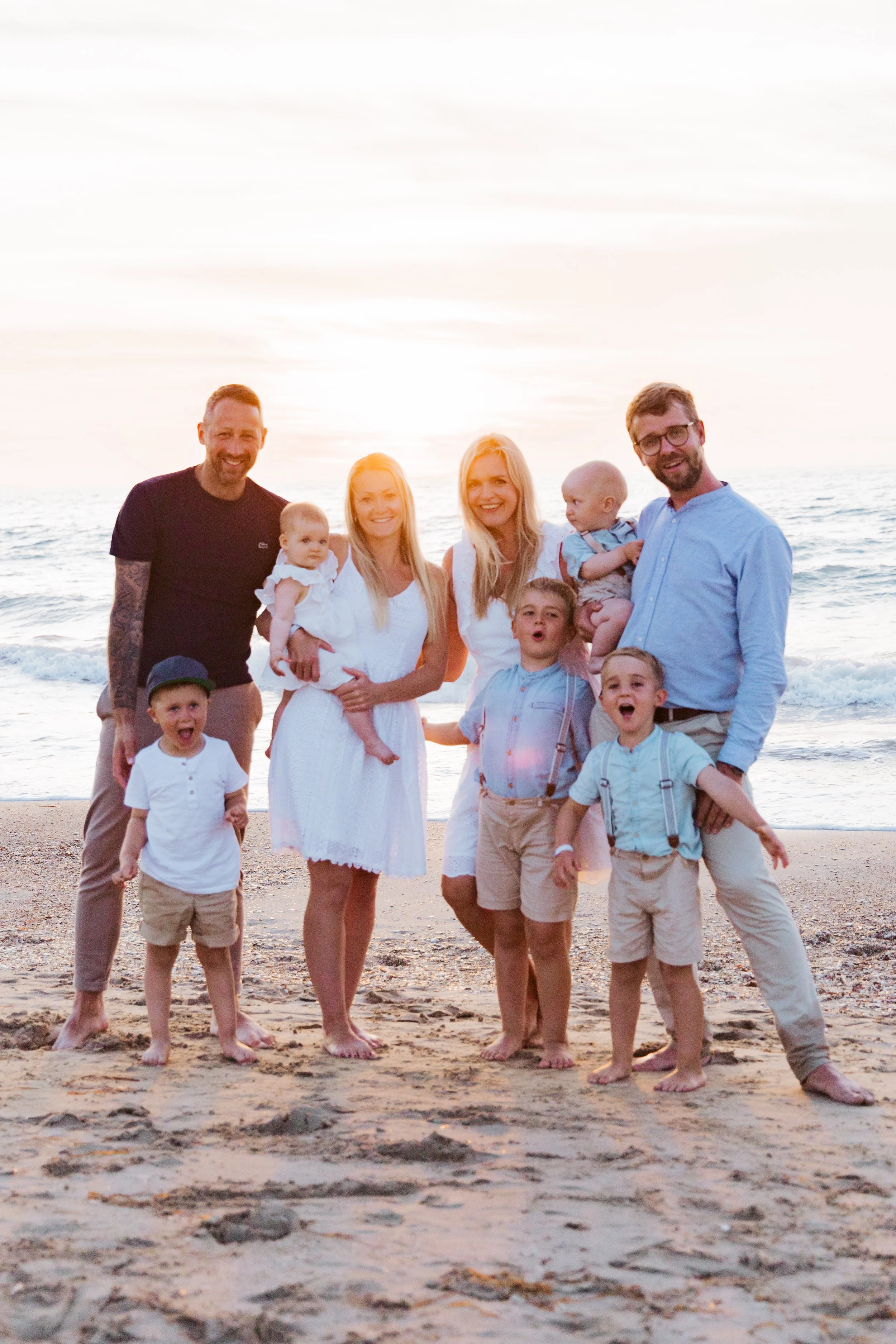 A happy family gathering on the beach at sunset, with adults and children smiling and playing near the water.