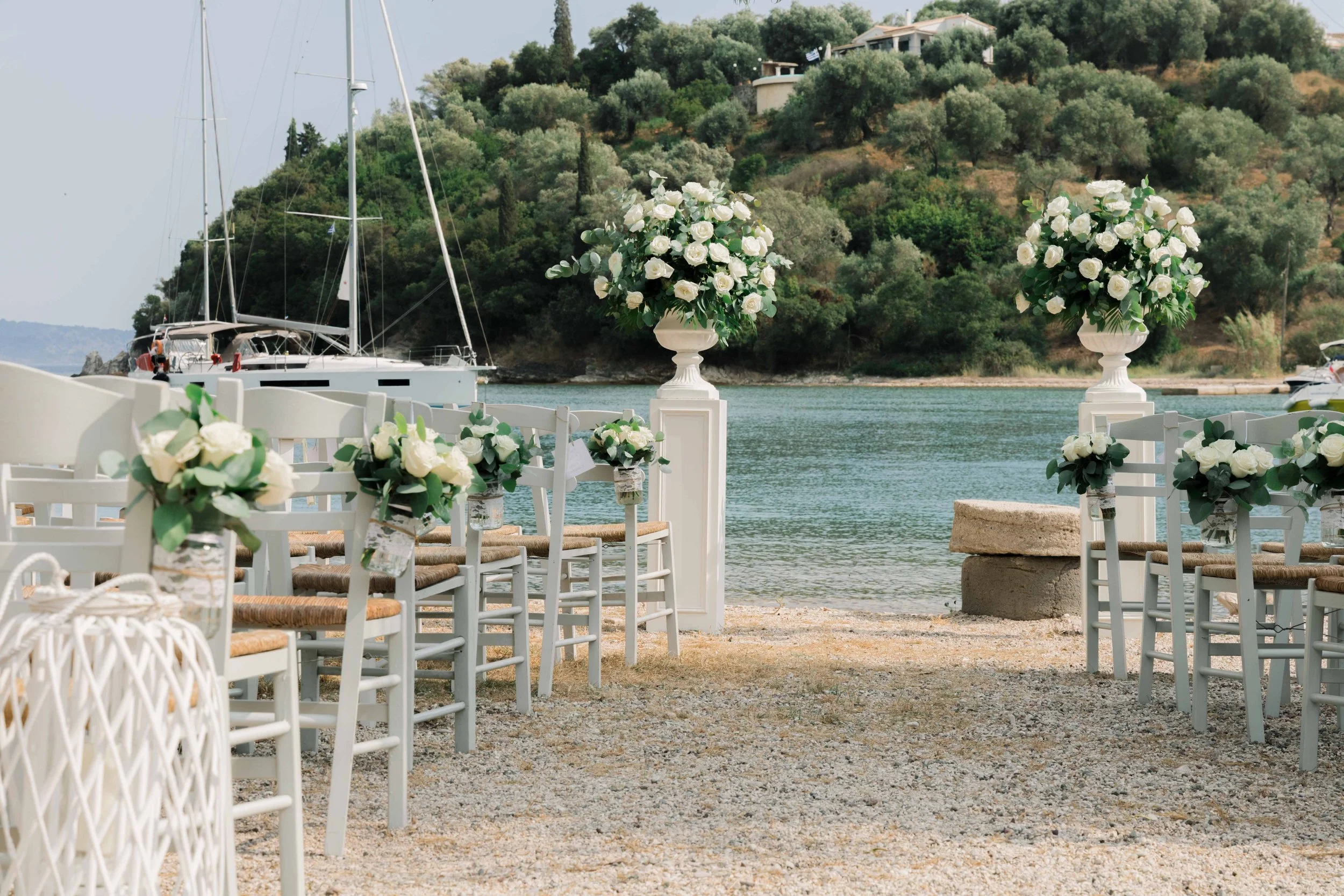 Outdoor wedding setup by the water with white chairs decorated with flowers, large floral arrangements on pedestals, and yachts docked in the background.