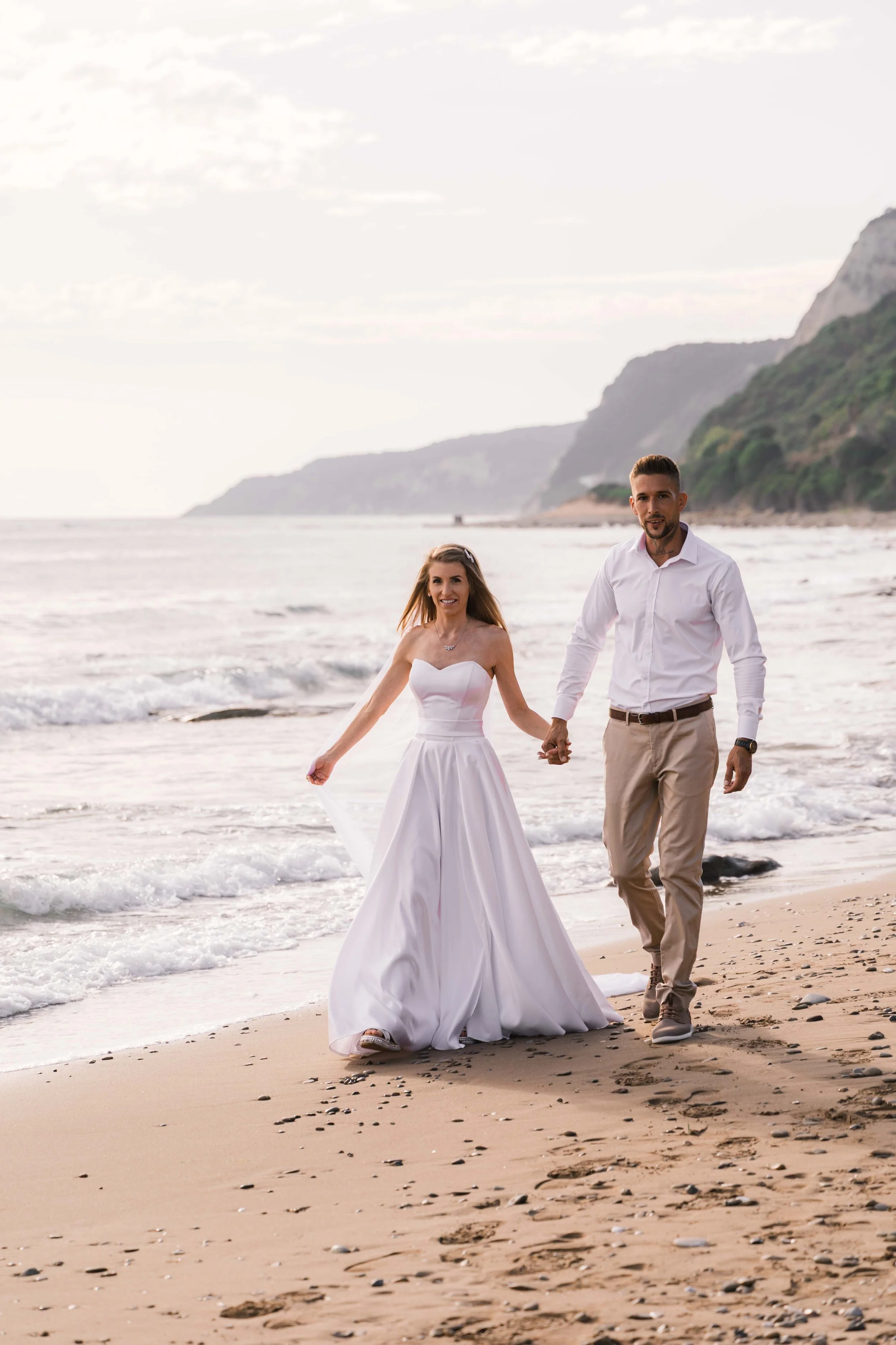 A couple in wedding attire walking hand in hand on a beach with the ocean and cliffs in the background.