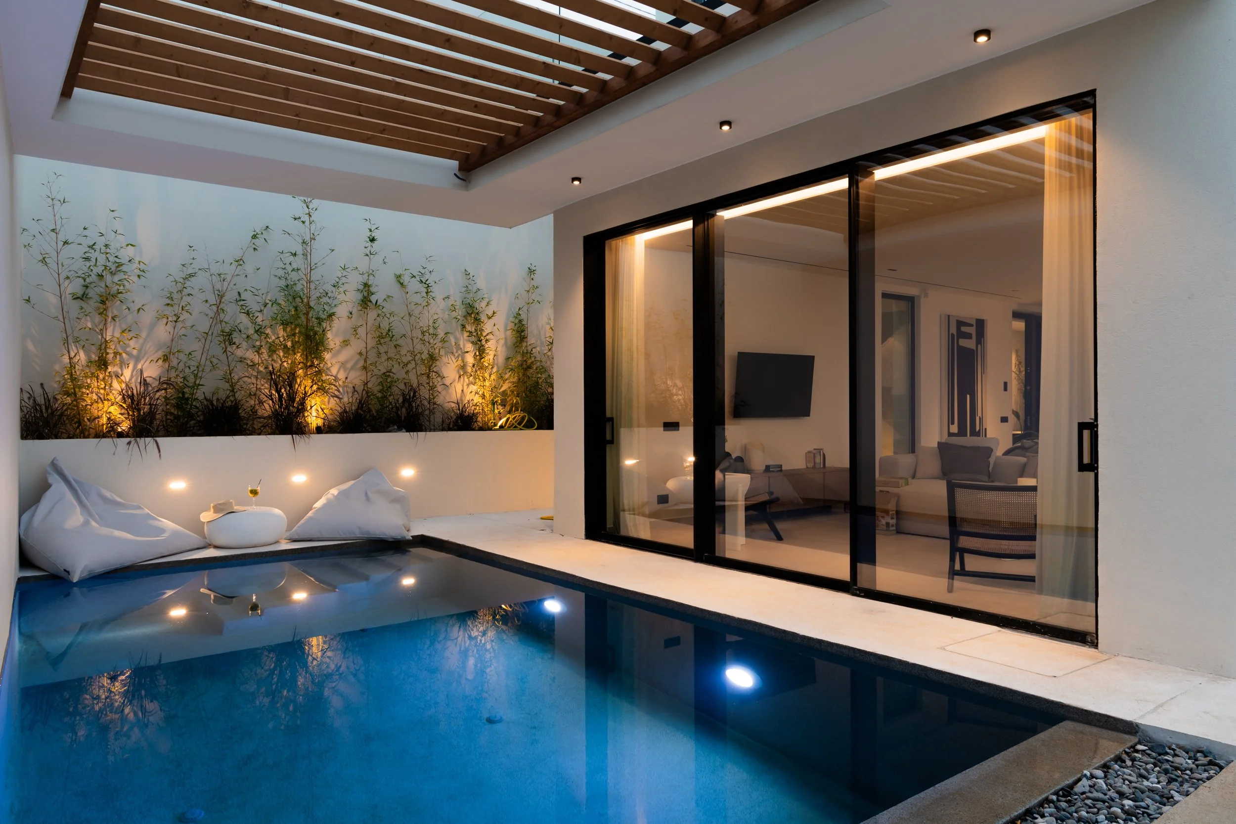 A modern house with a small outdoor swimming pool, glass sliding doors, and illuminated plants and seating area
