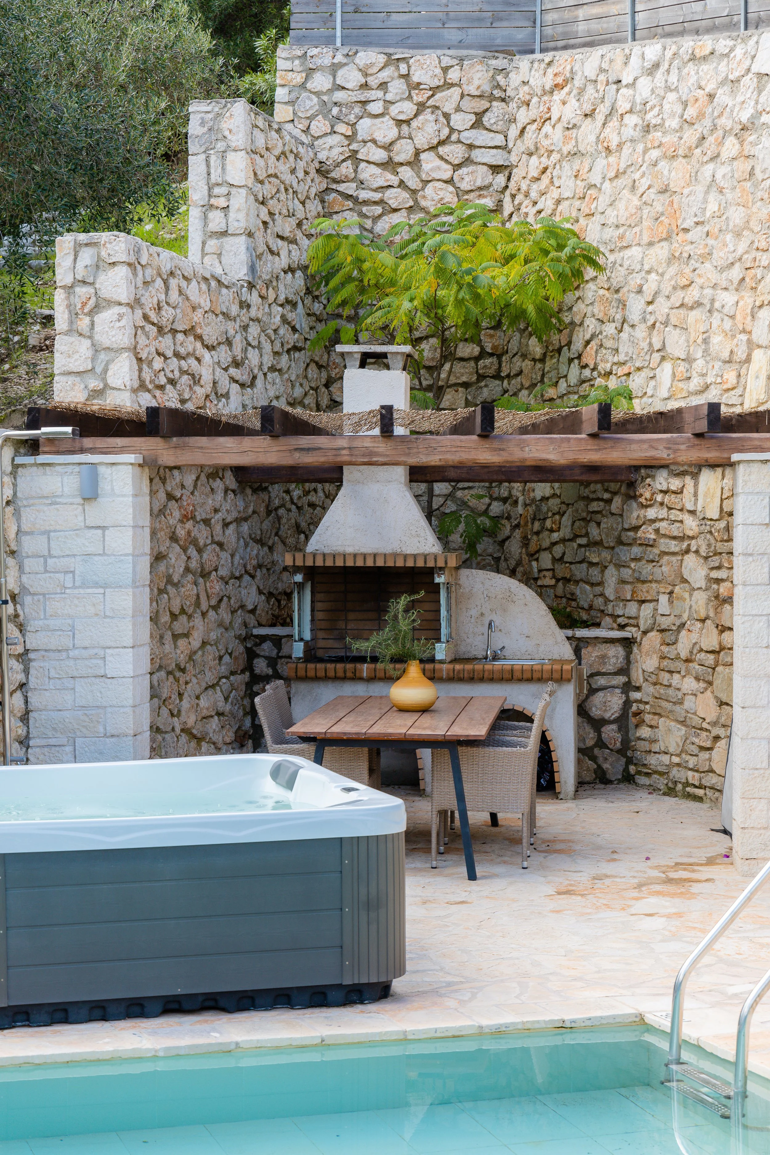A backyard scene featuring a hot tub in the foreground, a wooden dining table with four chairs and a decorative vase with greenery, a built-in brick grill, and stone walls with a small tree growing behind.