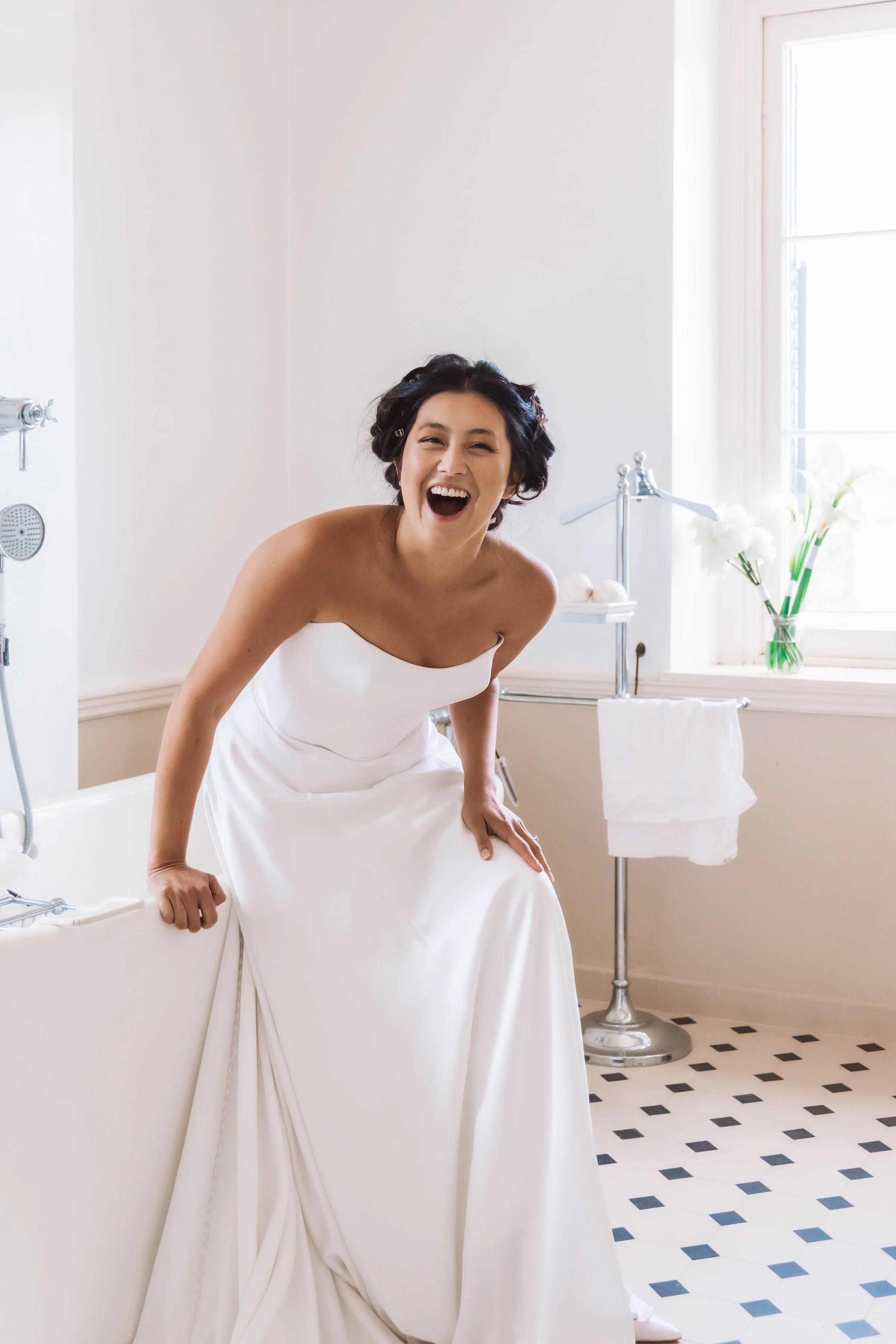 A woman in a white wedding dress sitting on the edge of a bathtub, laughing in a bright, airy bathroom with black and white tile floor and a vase of white flowers on the windowsill.