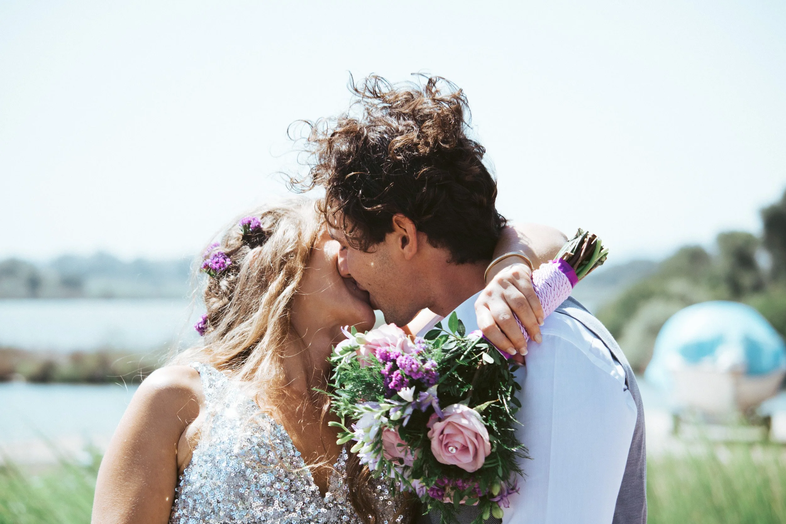 A couple kissing outdoors on their wedding day, with the bride holding a bouquet of pink and purple flowers, and the groom in a vest and dress shirt. The bride has long, wavy blonde hair with small purple flowers.