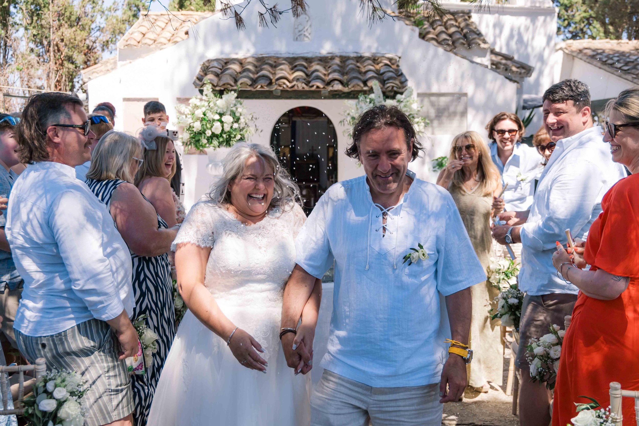 A wedding celebration outside with a bride and groom walking hand in hand while guests cheer and throw confetti in front of a white building with a tiled roof.