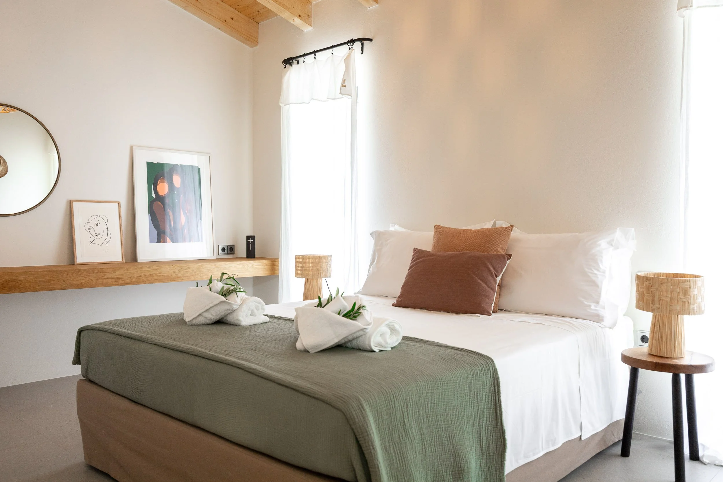 A cozy bedroom with a large bed decorated with white, brown, and beige pillows, a green bedspread, and towel swans with greenery. There are two wooden nightstands with woven lamps, framed artwork and a round mirror on the wall, and a window with whit