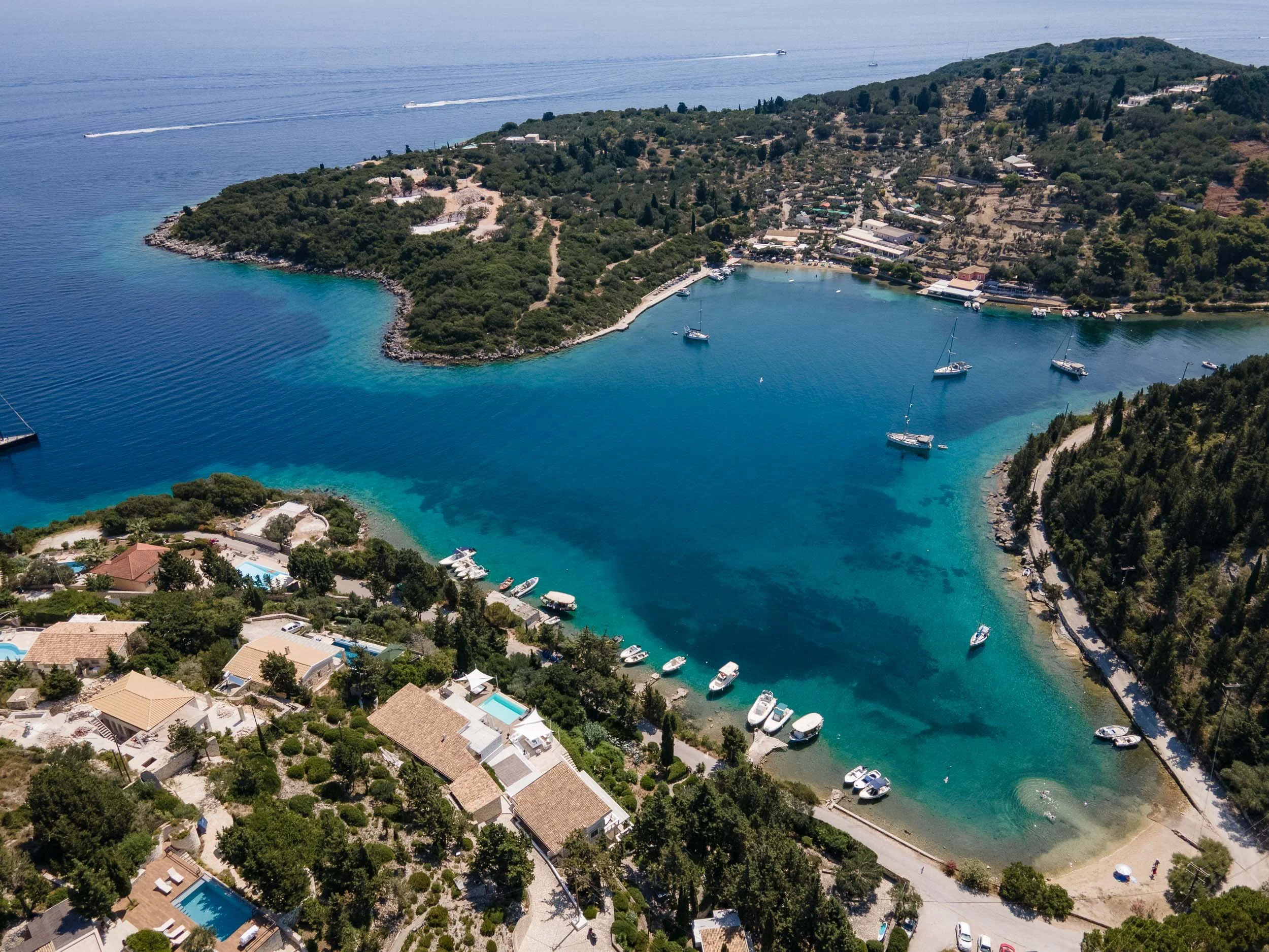Aerial view of a coastal residential area with boats anchored in a lagoon and houses with pools surrounded by trees.