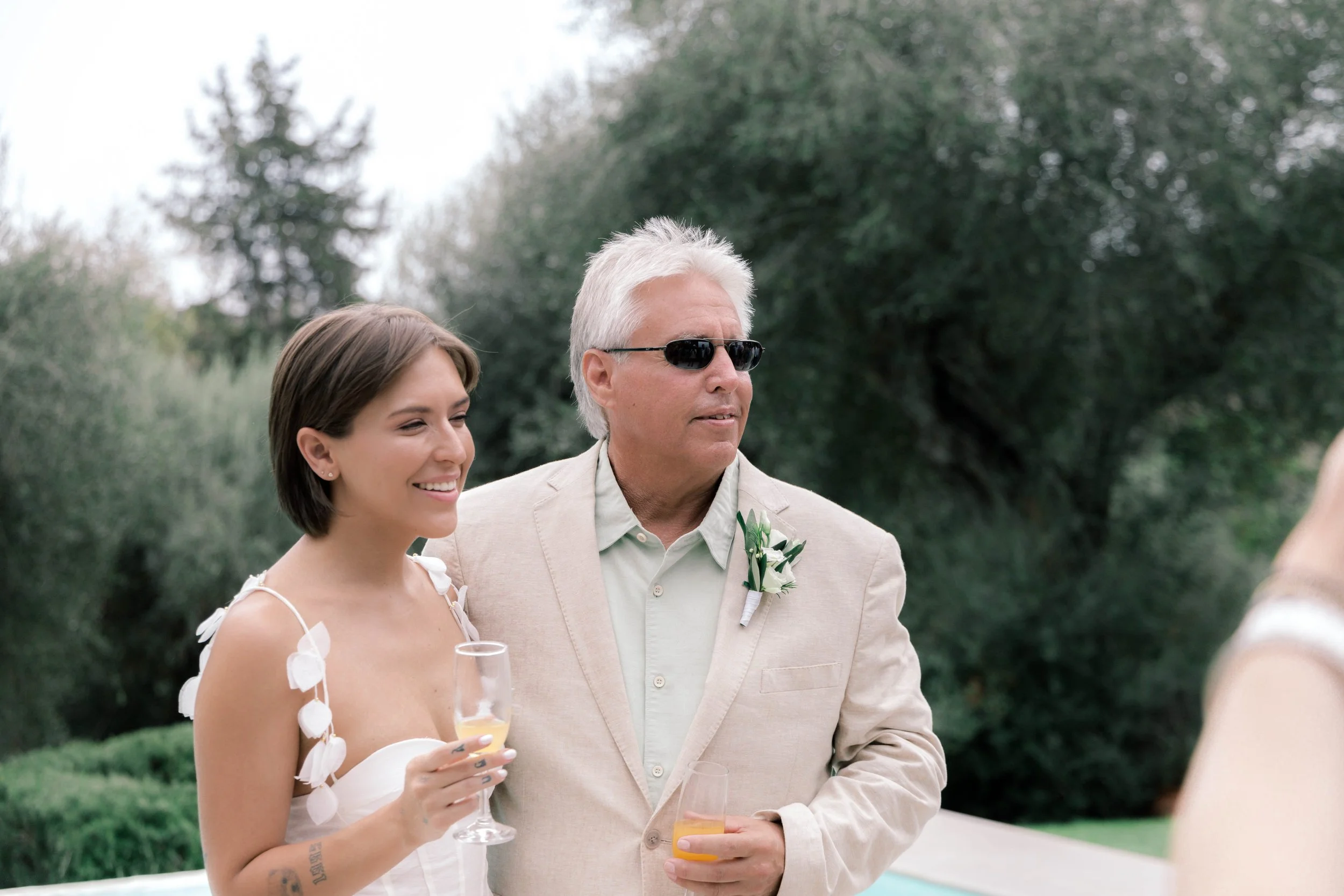 A woman in a white wedding dress holding a champagne glass, standing next to an older man in a beige suit and sunglasses, during an outdoor wedding celebration with greenery in the background.