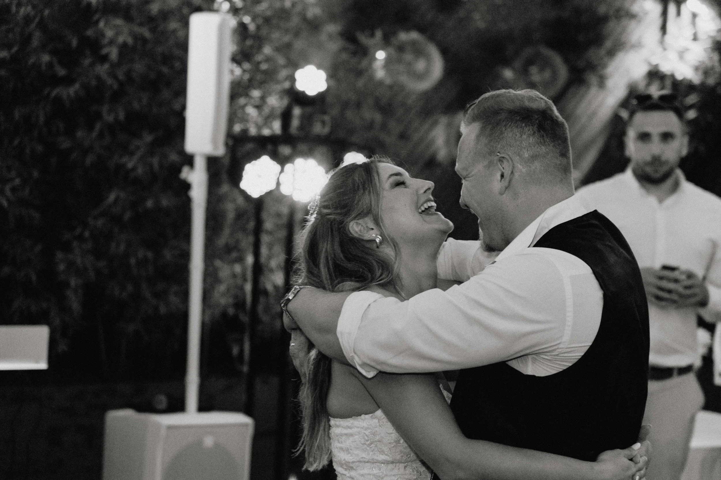 A joyful couple dancing and laughing together at a celebration outdoors, with another man in the background watching them.