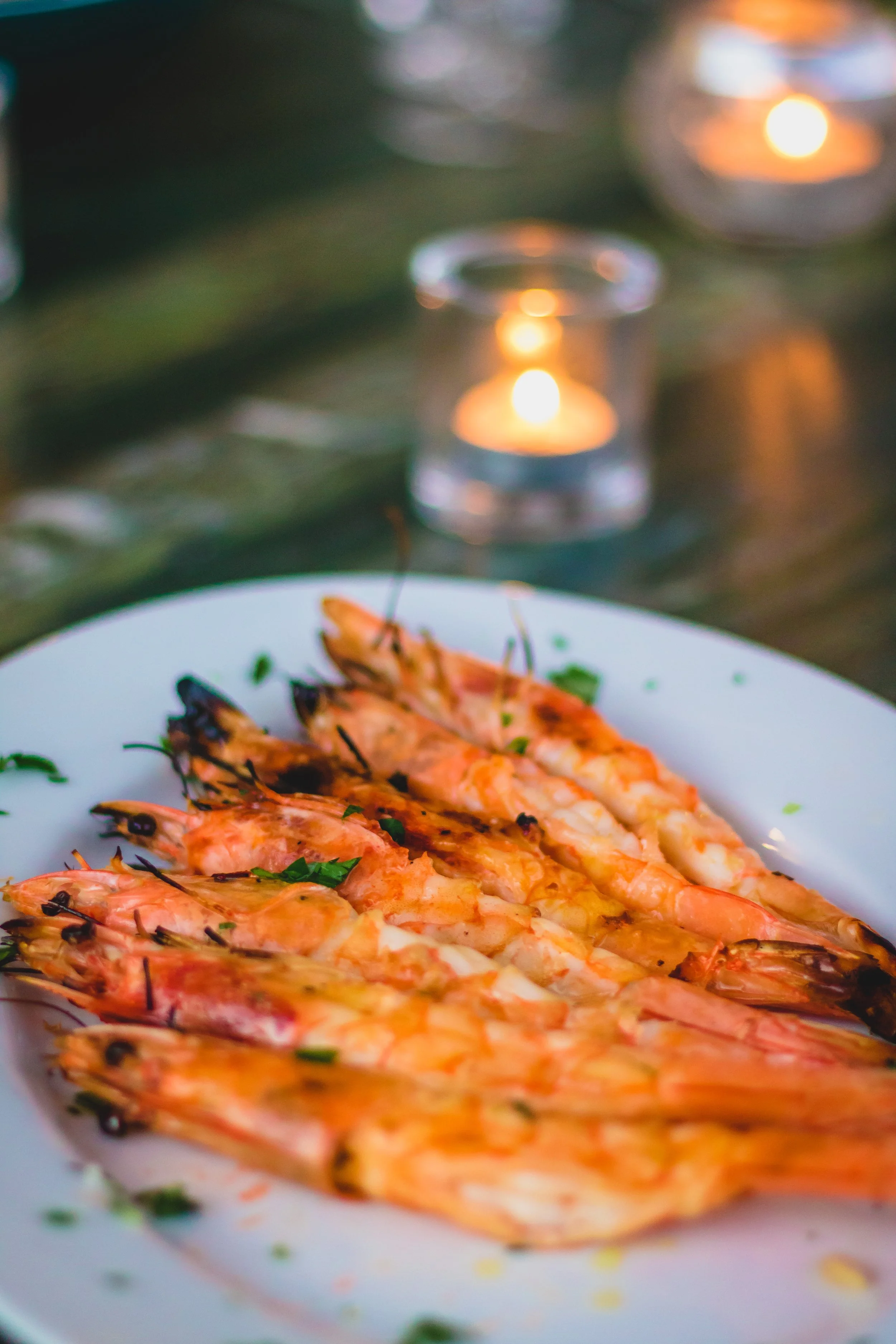 Plate of grilled shrimp garnished with herbs and two small candles in the background.