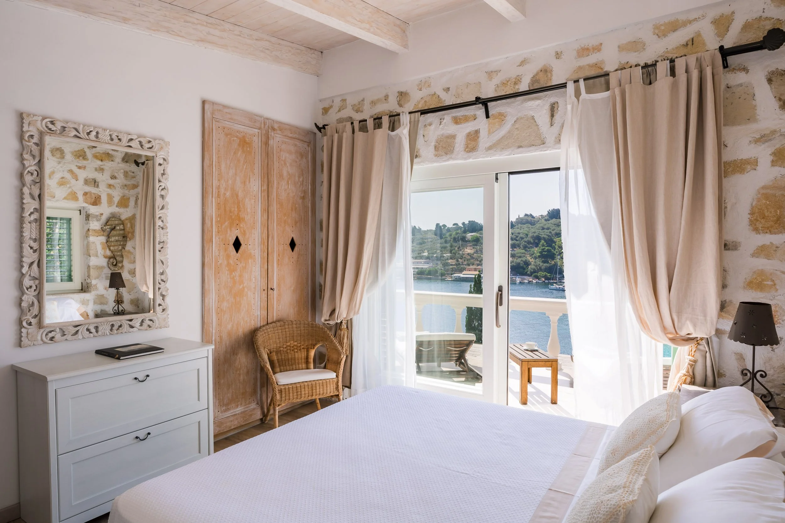 A cozy bedroom with white walls, a large window with beige curtains overlooking a river and green hills, a white dresser with a mirror, a wicker chair, and a bed with white linens.