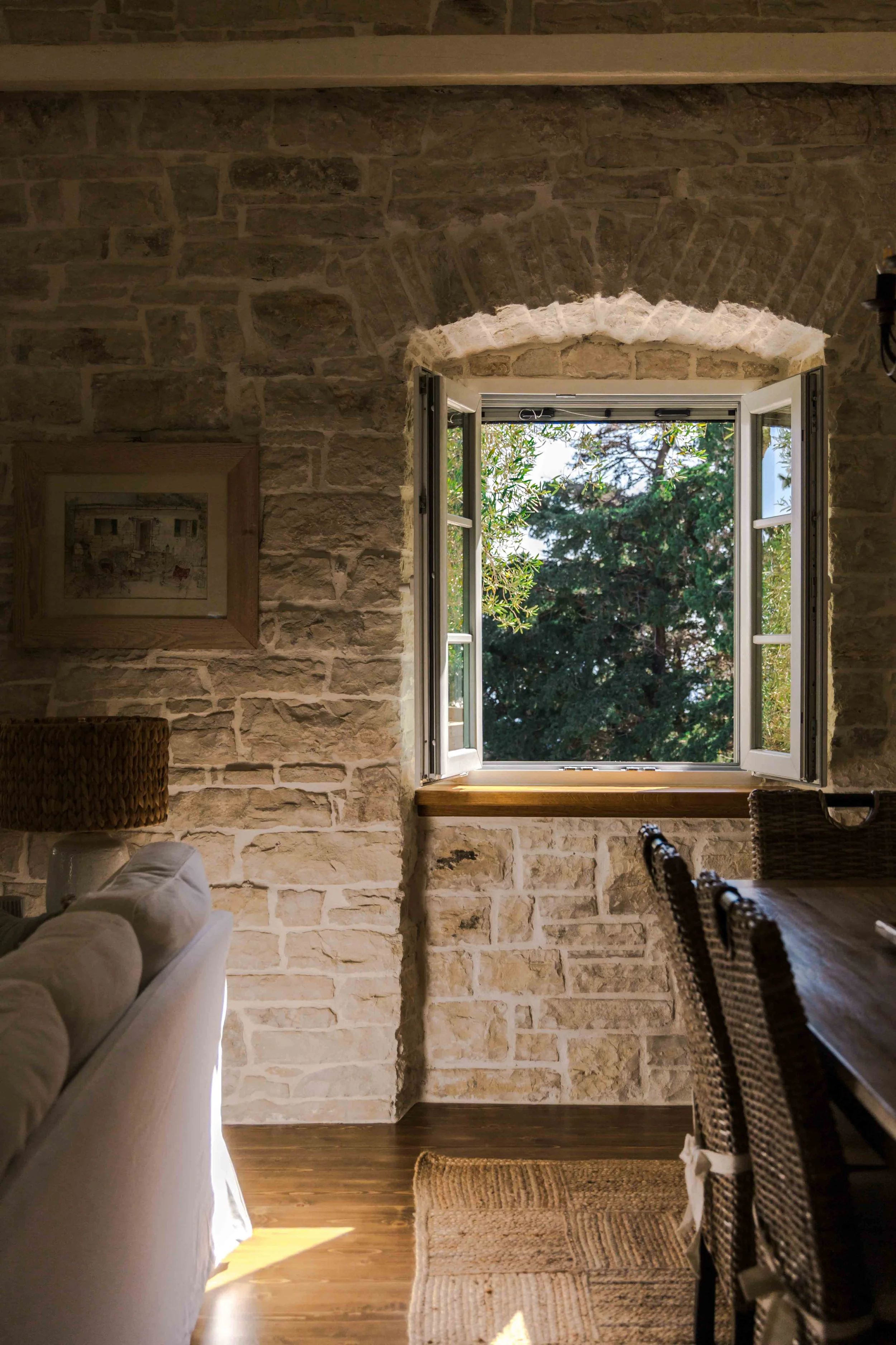 A cozy interior room with a stone wall, an open window showing trees outside, a sofa, a woven lamp, a painting on the wall, and a dining table with chairs, illuminated by natural light.