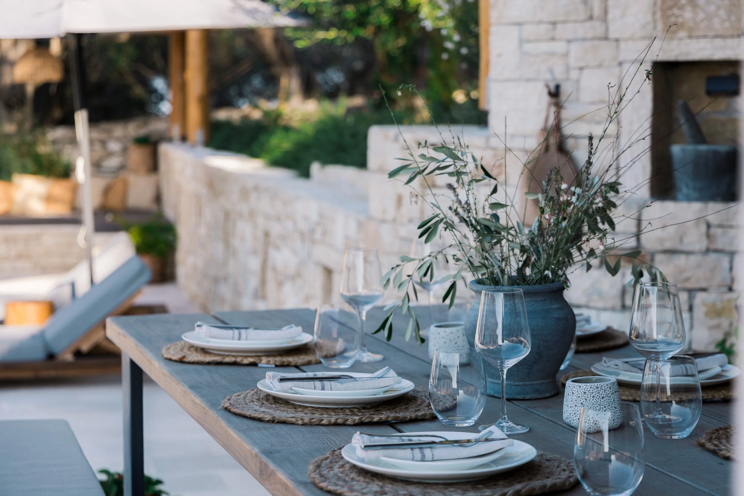 Outdoor dining table set with plates, glasses, utensils, woven placemats, and a large vase with greenery, on a patio with stone wall and greenery in background.