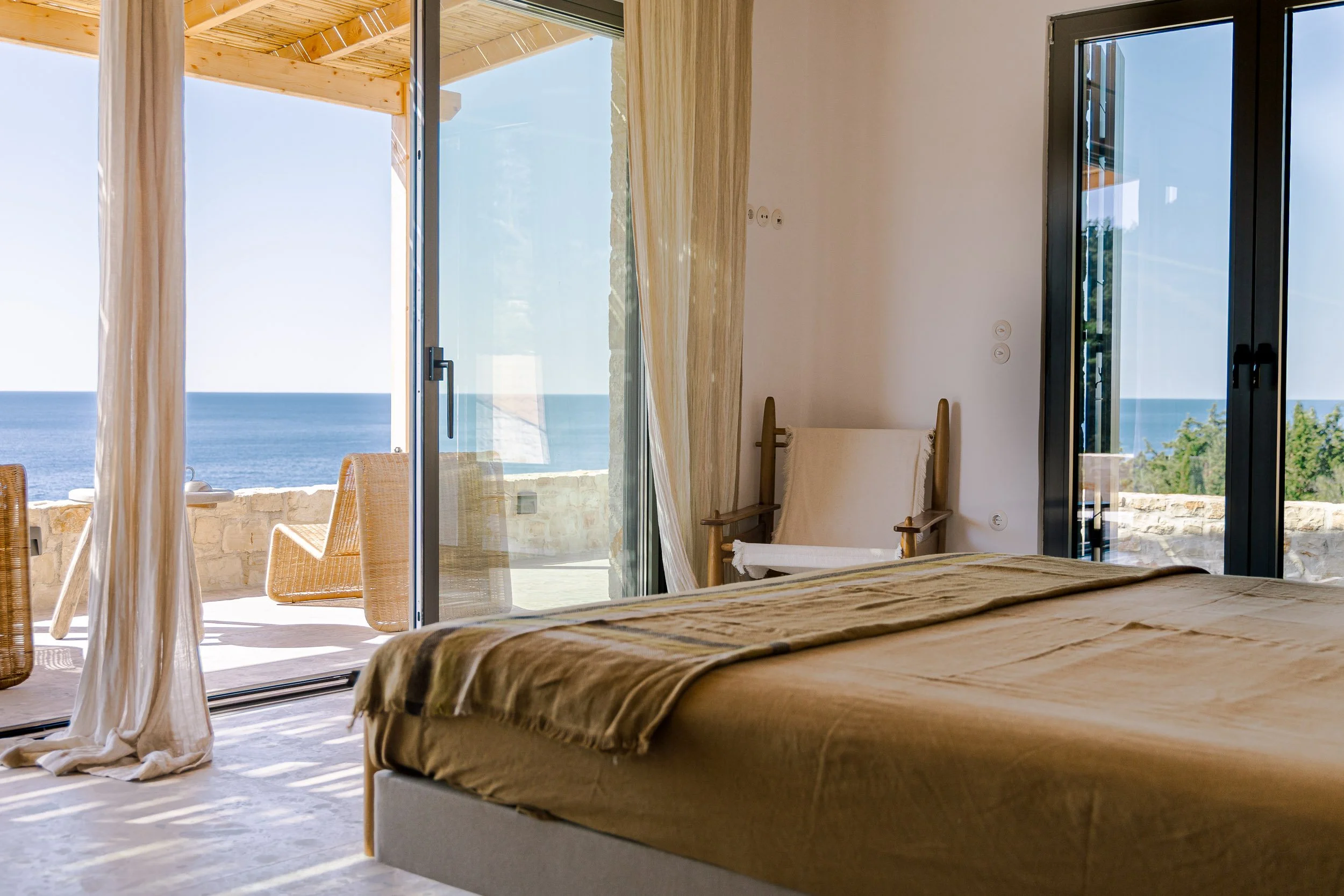 A bedroom with a bed covered in tan bedding, open glass doors leading to a balcony with ocean view, and a wooden chair with a beige cushion.