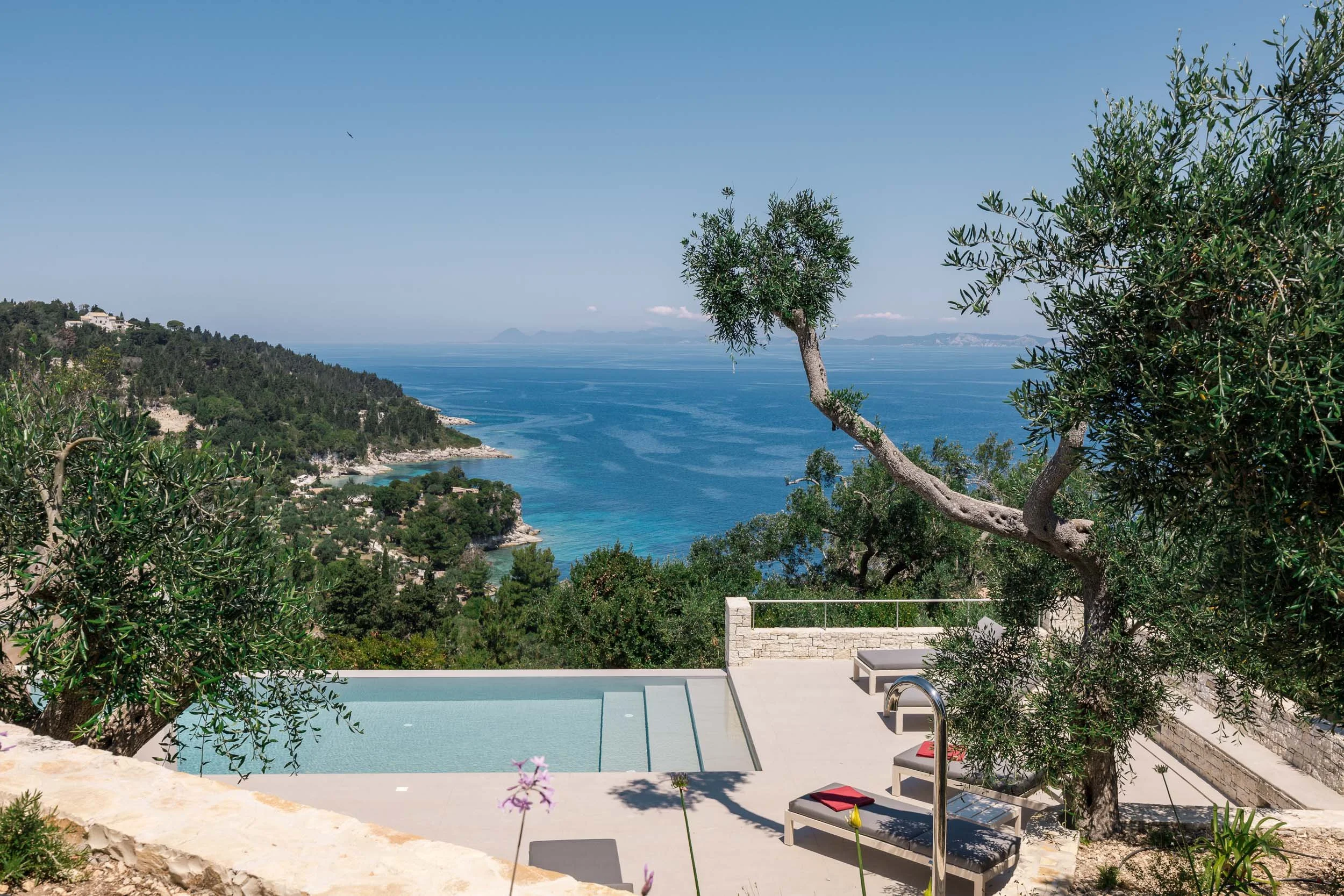 A scenic coastal view with an infinity pool, lounge chairs with red pillows, and trees on a terrace overlooking a blue ocean with hills and islands in the distance.