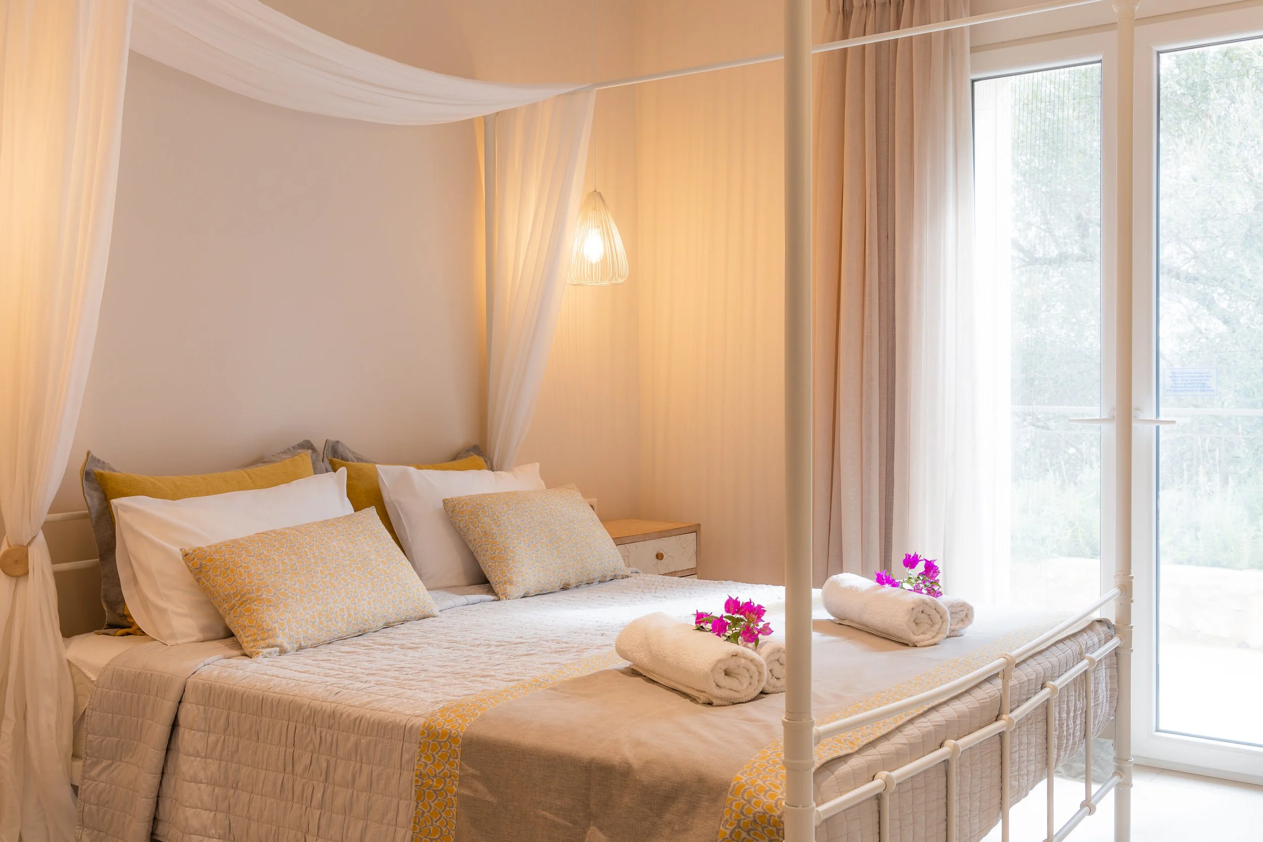 A cozy bedroom with a white four-poster bed with white, yellow, and gray pillows, rolled towels with pink flowers, sheer curtains, and a glass door leading outside.