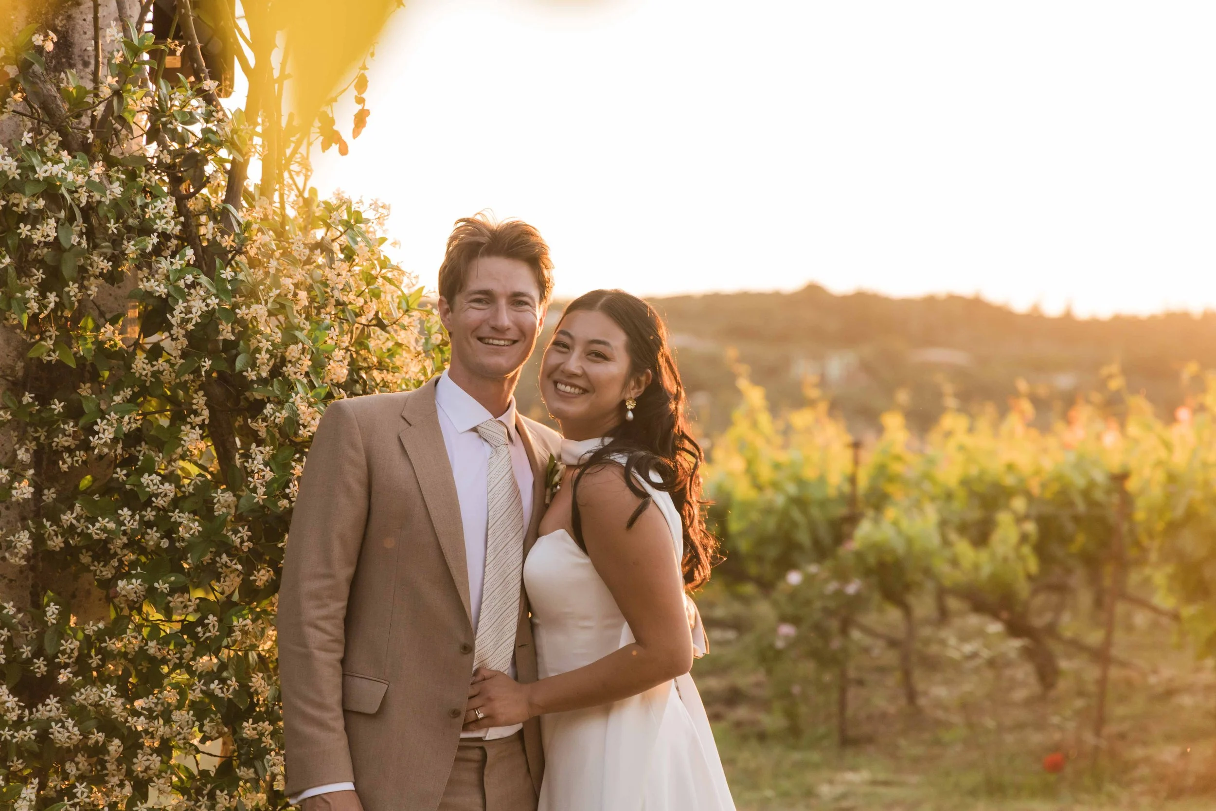 A smiling couple stands in a vineyard at sunset, with the man wearing a beige suit and the woman in a white dress.