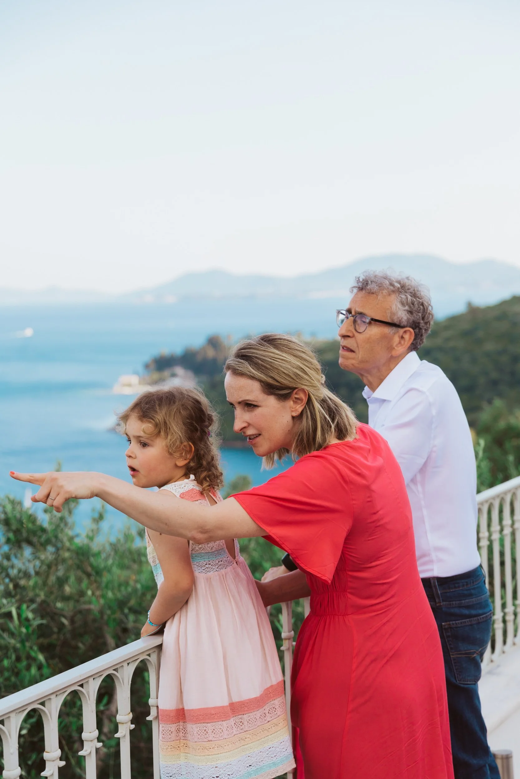A woman, a young girl, and an older man are standing on a balcony overlooking the water and mountains in the background. The woman is pointing at something in the distance.