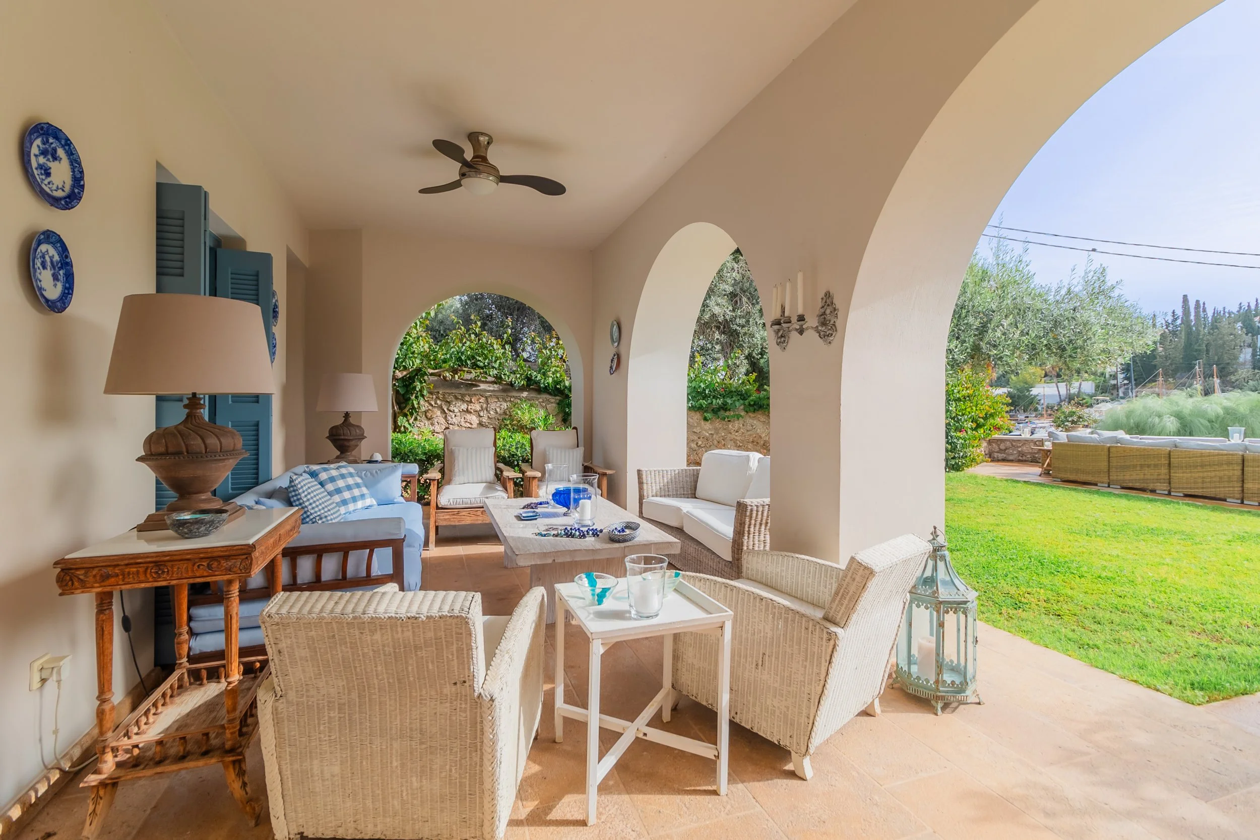 Covered patio with wicker and wooden outdoor furniture, including chairs, sofas, and a coffee table, overlooking a lush yard with green grass and trees.