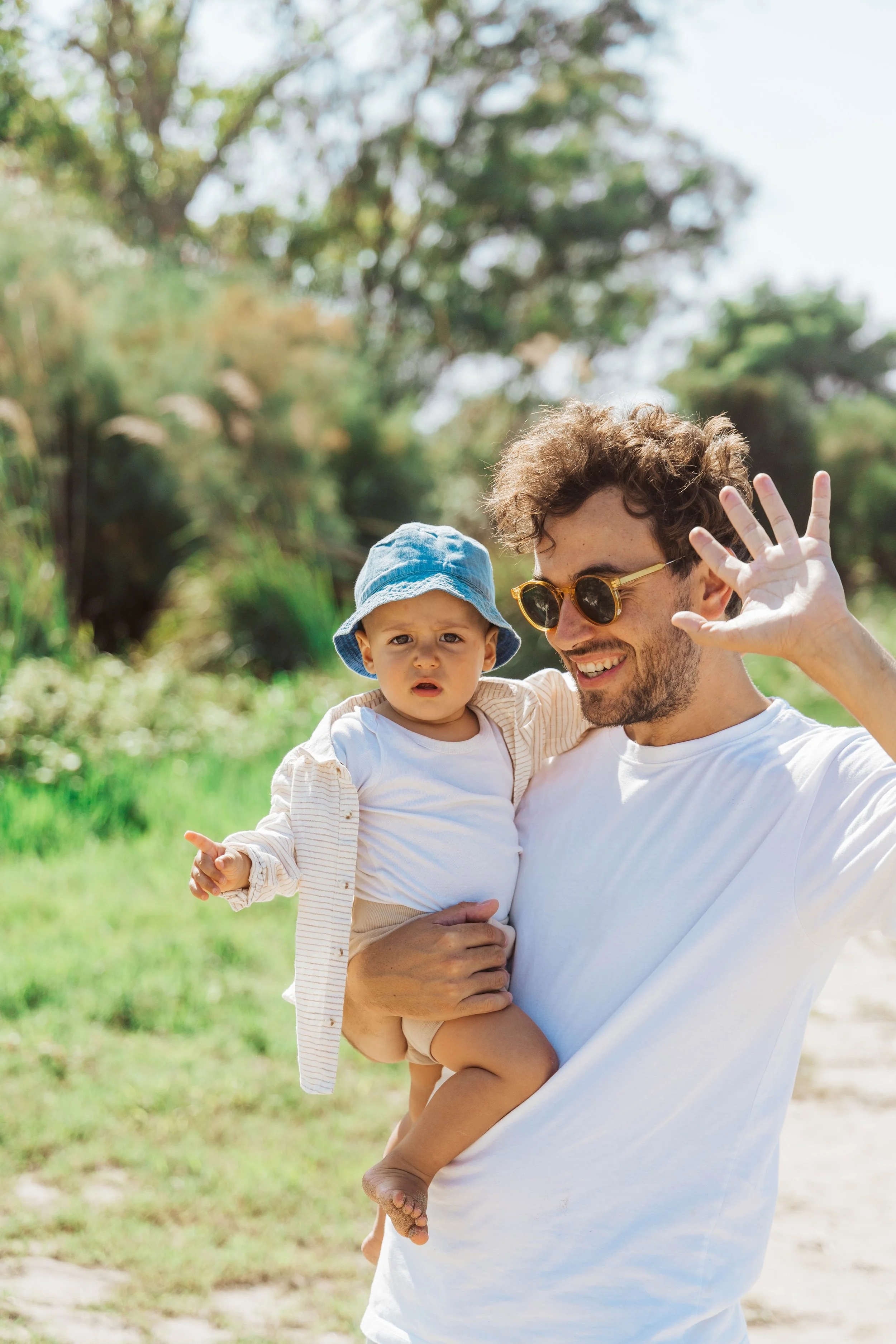 A man wearing sunglasses is smiling and waving while holding a young child in his arms outdoors in a park or natural setting.