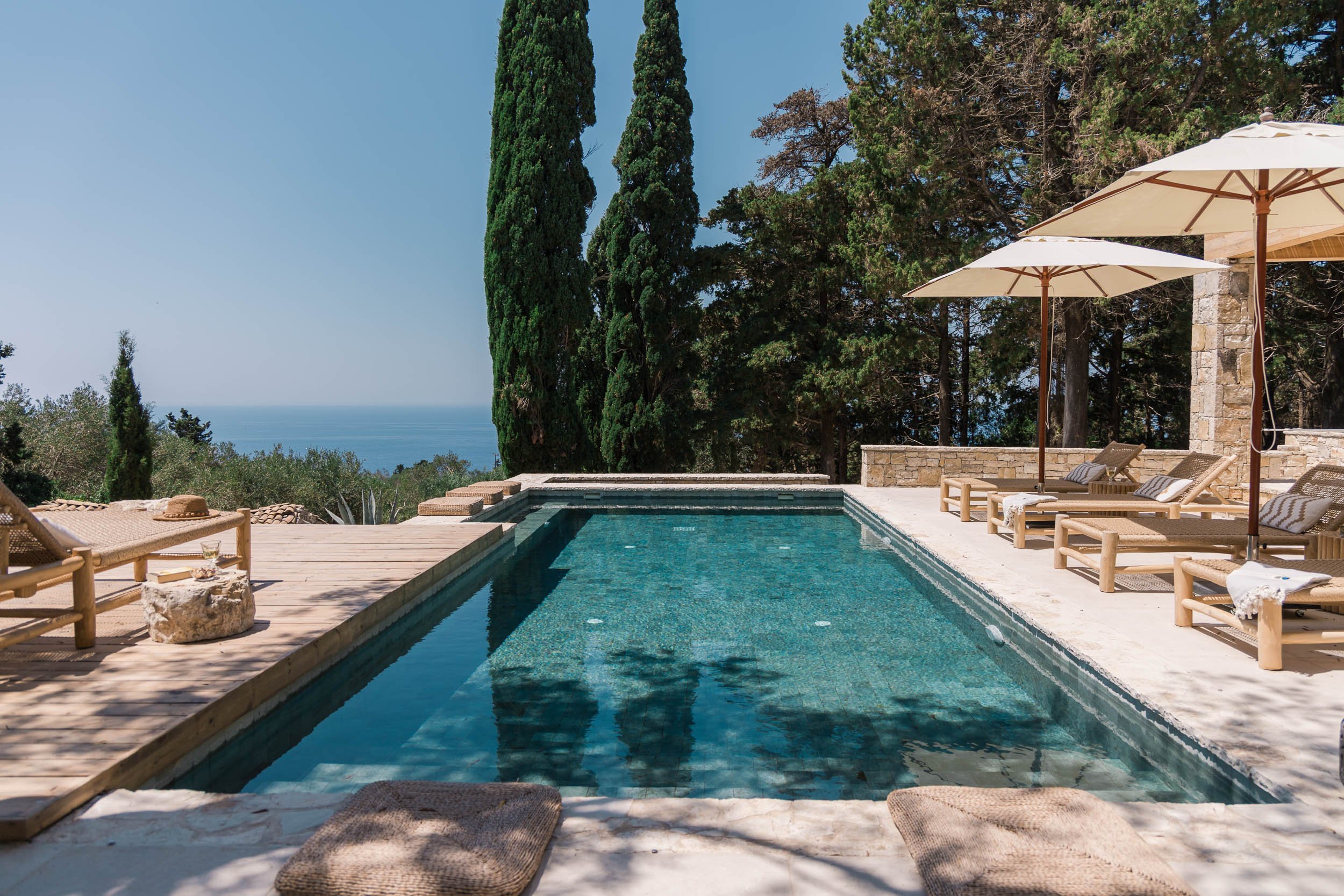 View of a luxurious swimming pool with lounge chairs and umbrellas on a wooden pool deck, surrounded by tall trees overlooking the ocean in the background.
