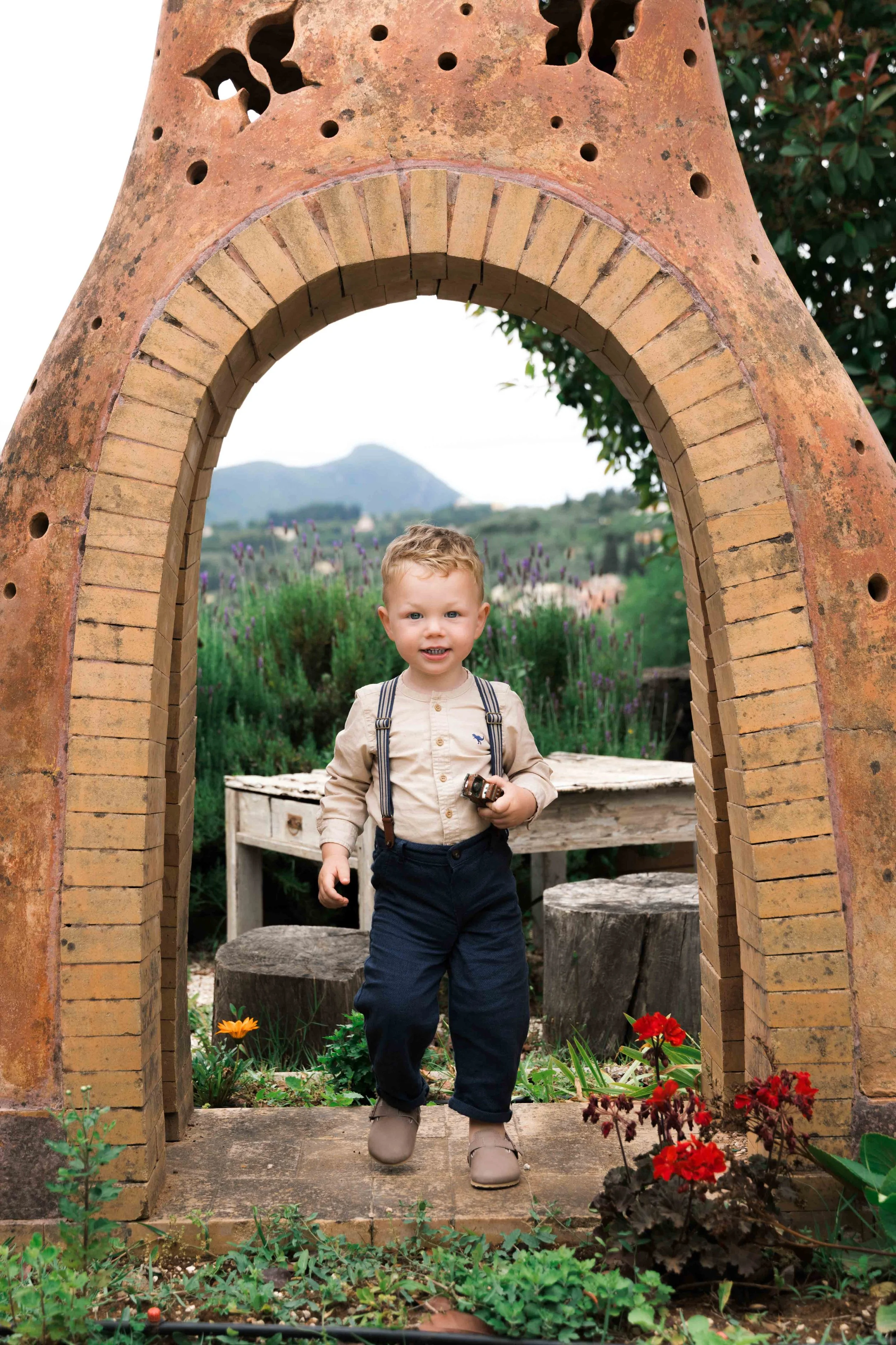 A young boy standing outside under a brick archway, holding a small toy, with trees, flowers, and mountains in the background.
