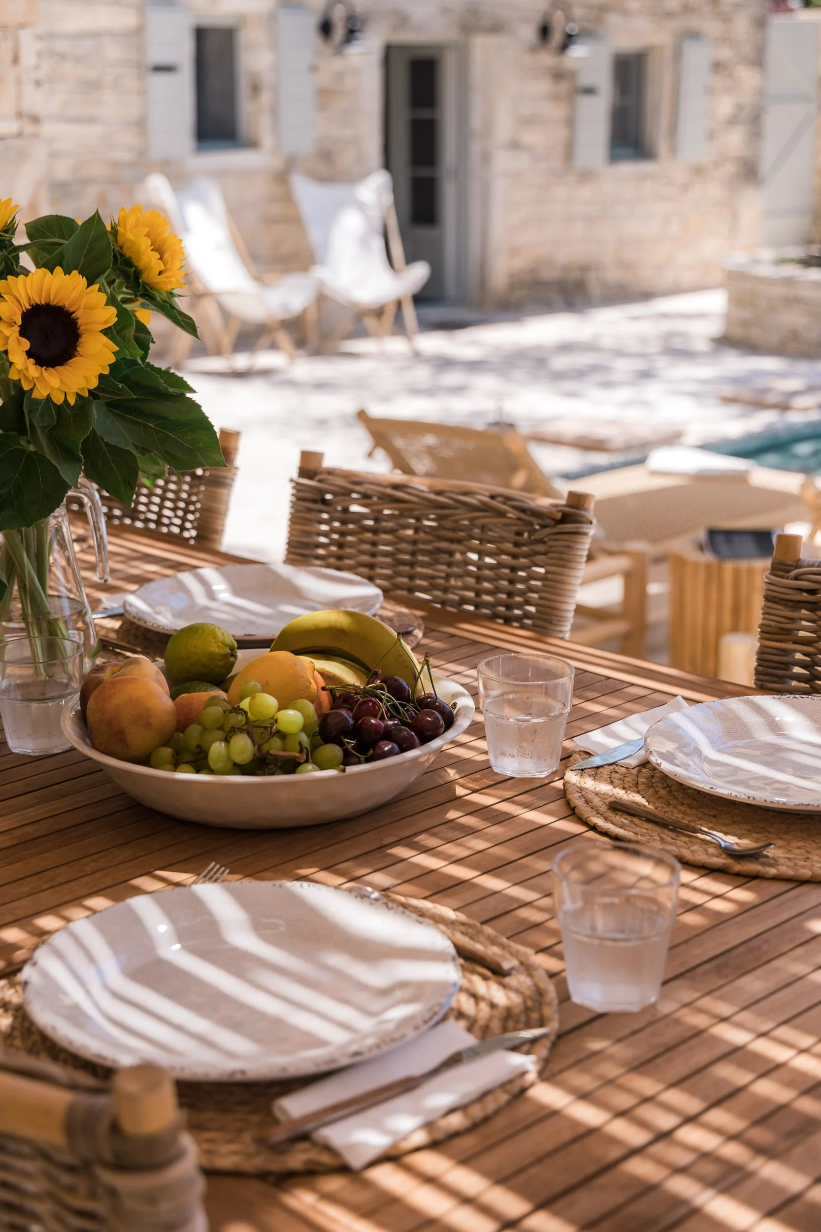 An outdoor dining table set with plates, cutlery, glasses of water, a bowl of assorted fruits including apples, bananas, grapes, and a lemon, and a sunflower centerpiece. The table is on a patio next to a stone building with lounge chairs nearby.