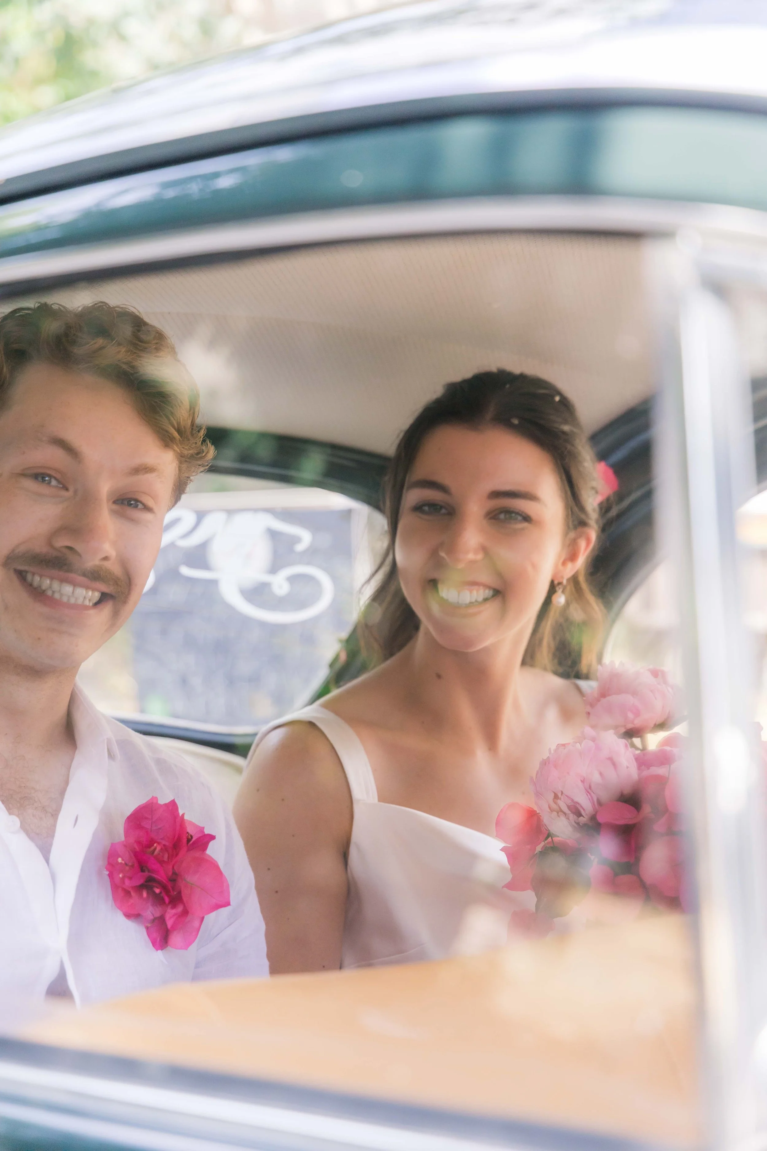 A smiling man and woman sitting inside a vintage car, holding pink flowers, during a wedding celebration.