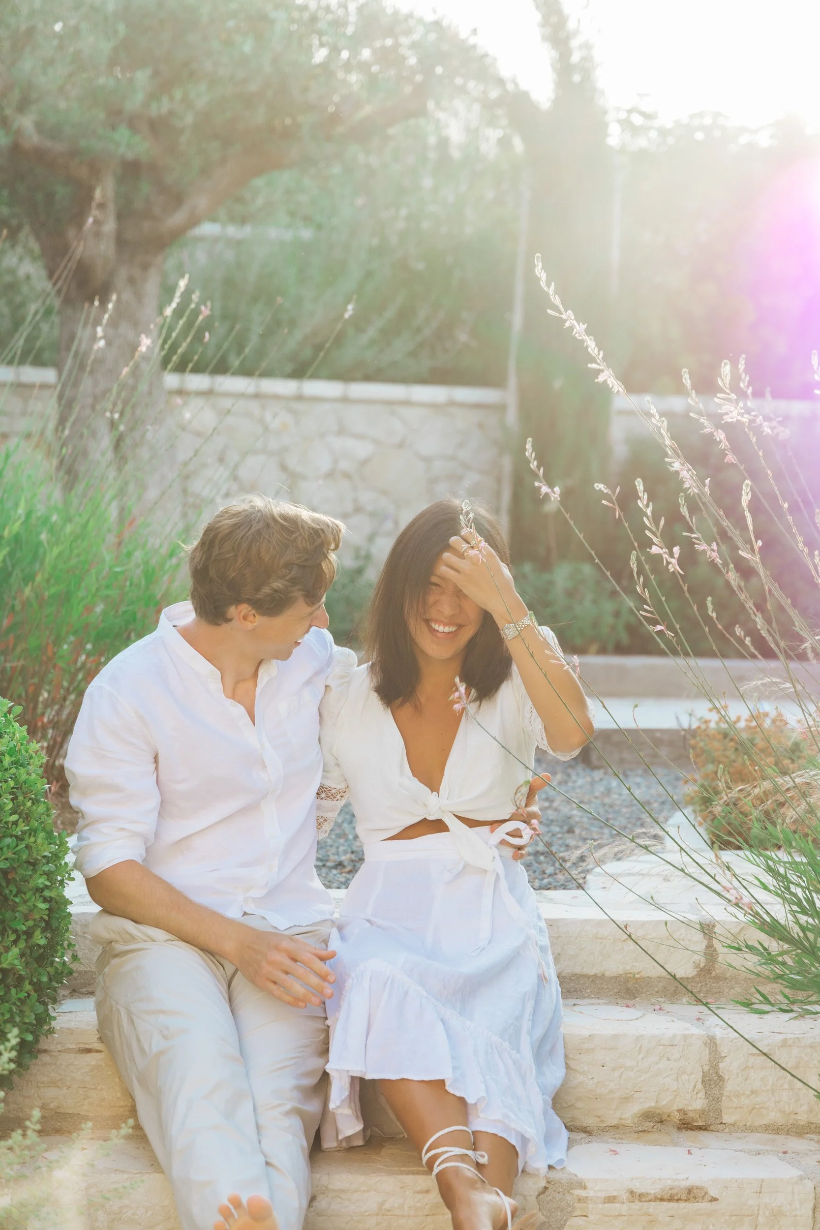 A young man and woman sitting on stone steps outdoors, smiling and enjoying each other's company with sunlight shining through nearby trees.