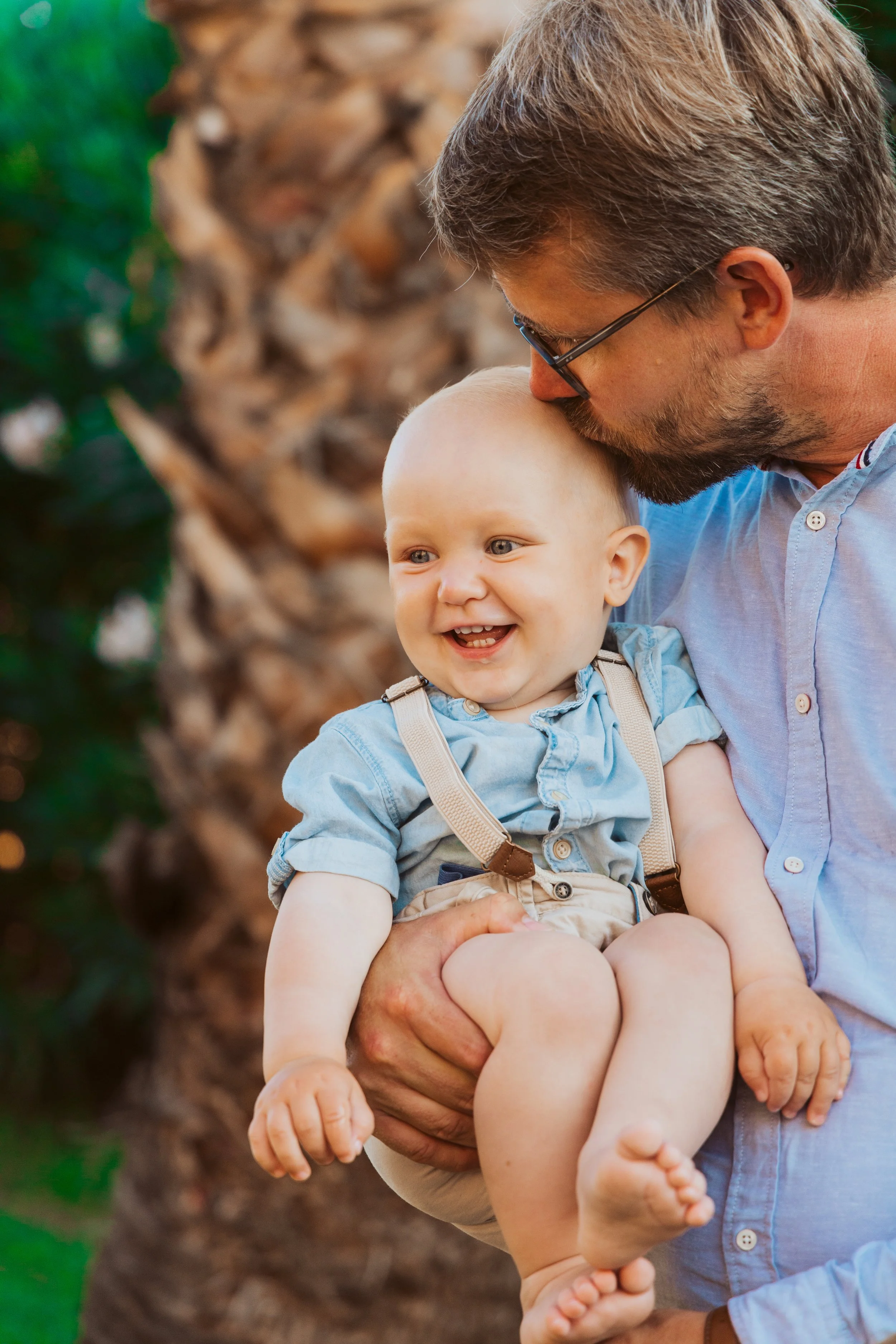 A man with glasses and a beard holding a smiling baby boy outdoors near a palm tree.