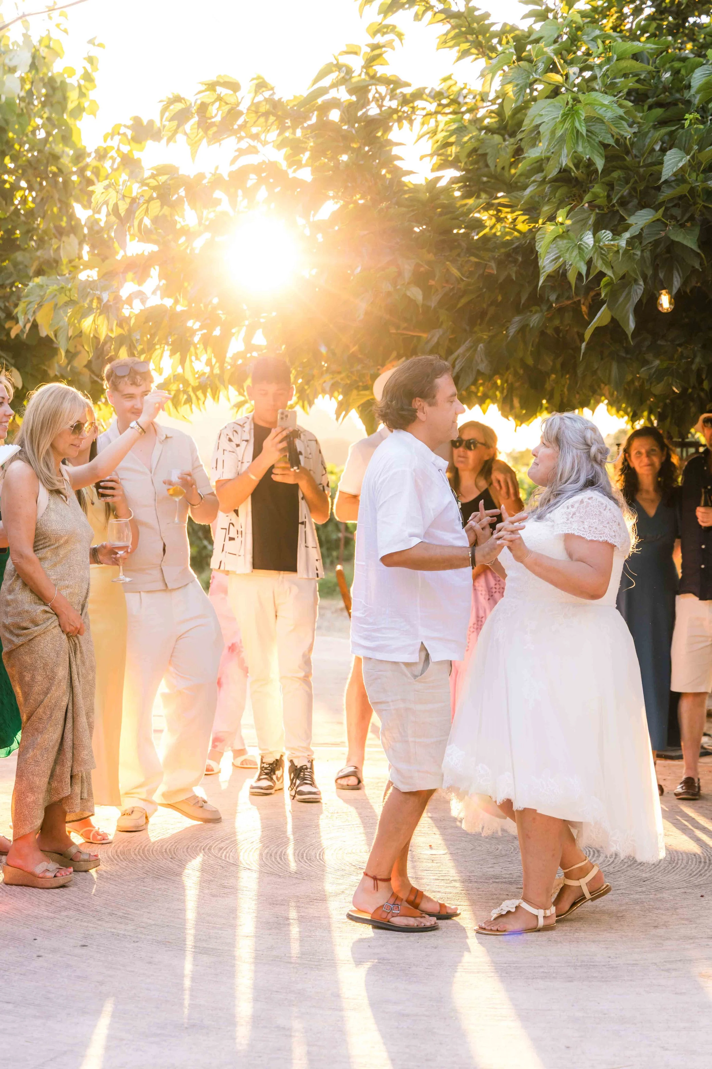 Couple dancing at a wedding reception during sunset, surrounded by friends holding drinks and taking photos.