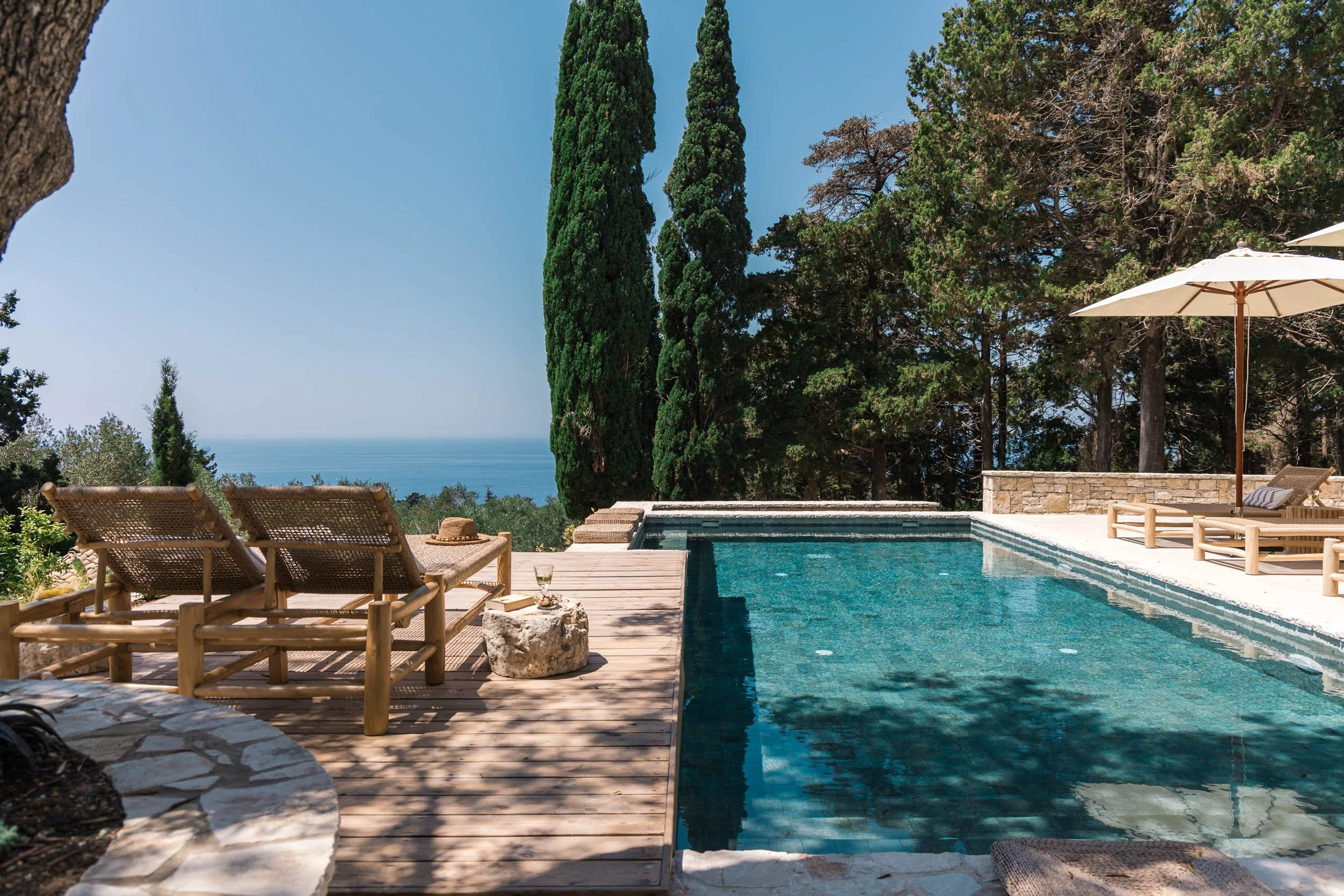 A backyard with a swimming pool surrounded by wooden deck chairs, umbrellas, and lush trees, overlooking the ocean in the distance.