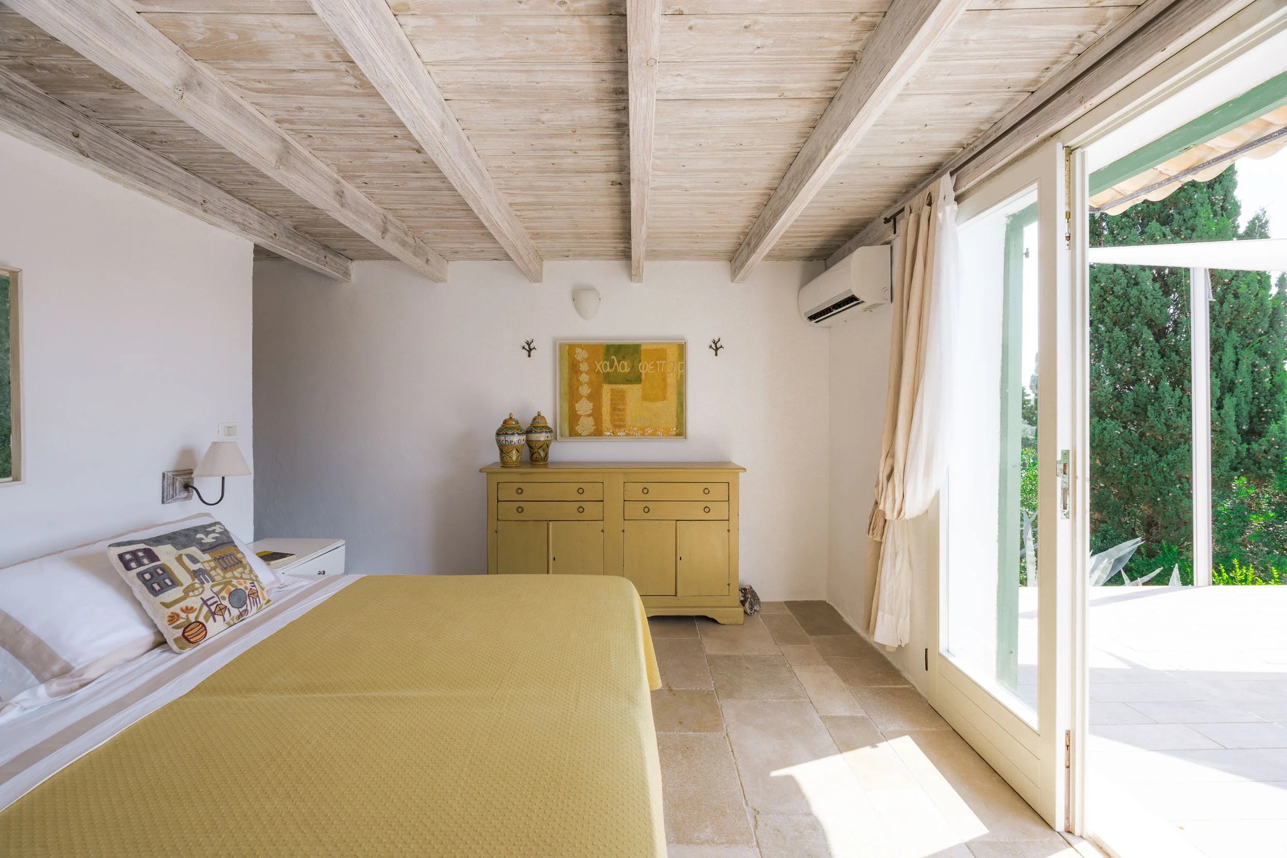 A bedroom with a yellow bedspread, a white pillow with decorative pillows, a wall-mounted lamp, a yellow dresser with decorative vases, framed wall art, and an open sliding glass door showing greenery outside.
