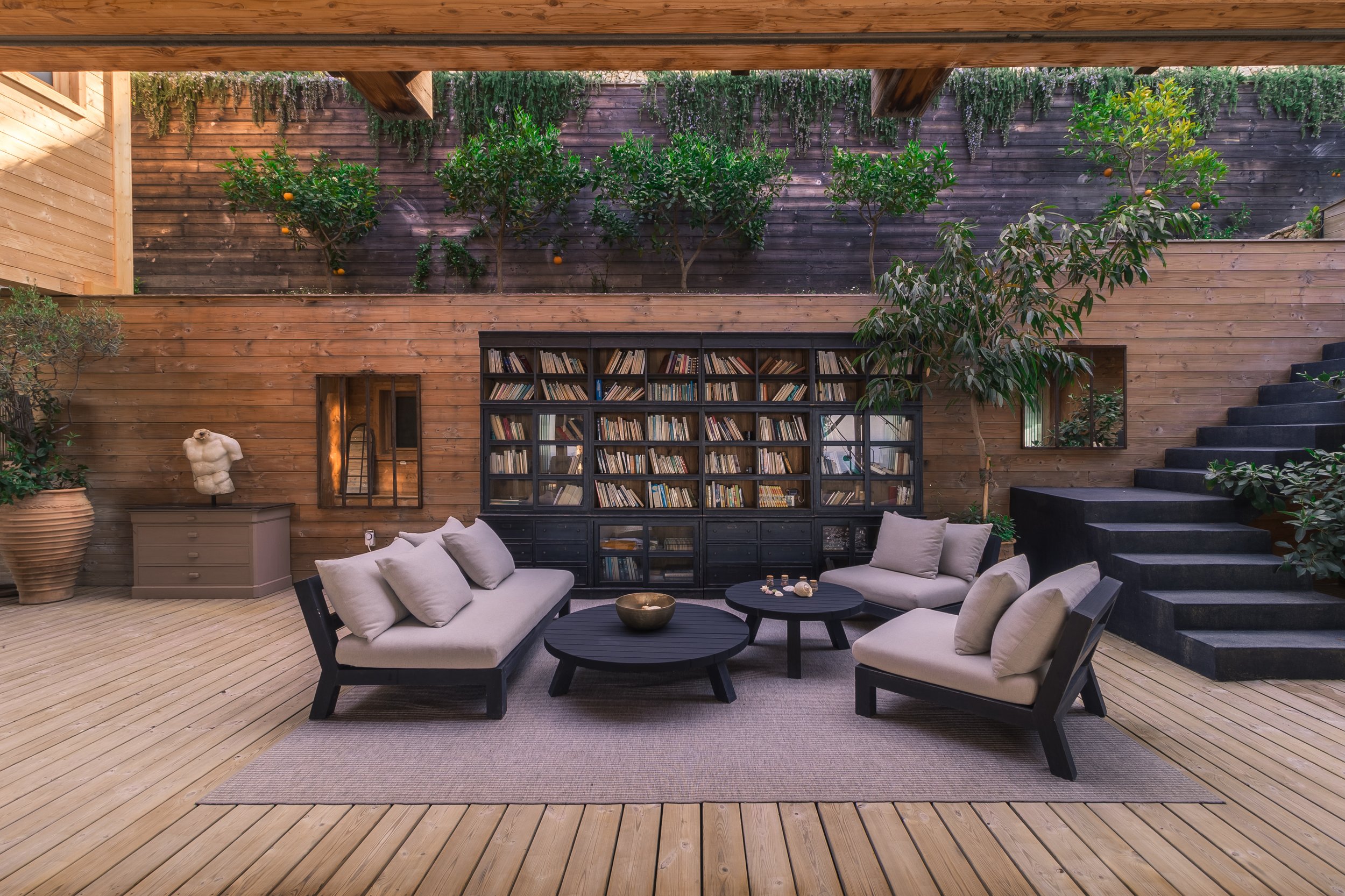Indoor patio with wooden walls and ceiling, black bookshelf filled with books, beige lounge furniture with pillows, black coffee tables with decorative items, potted plants, and a staircase on the right. Green trees are visible above the walls.