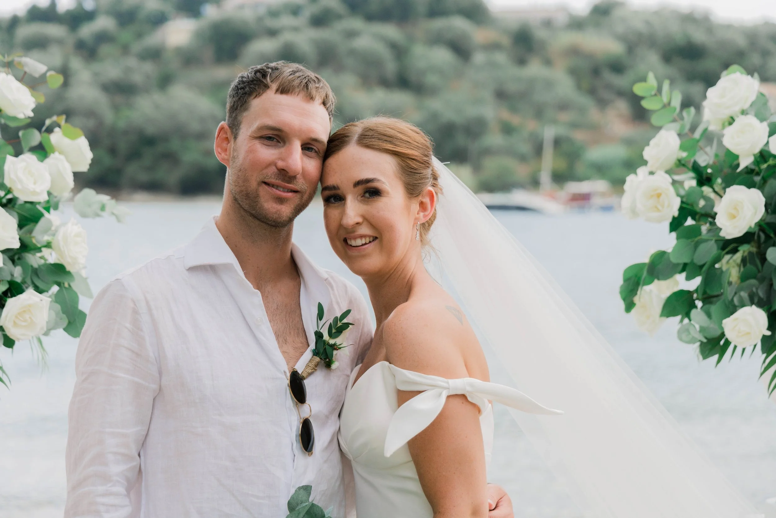 A bride and groom smiling together outdoors by a body of water, with greenery and boats in the background, surrounded by white floral arrangements.