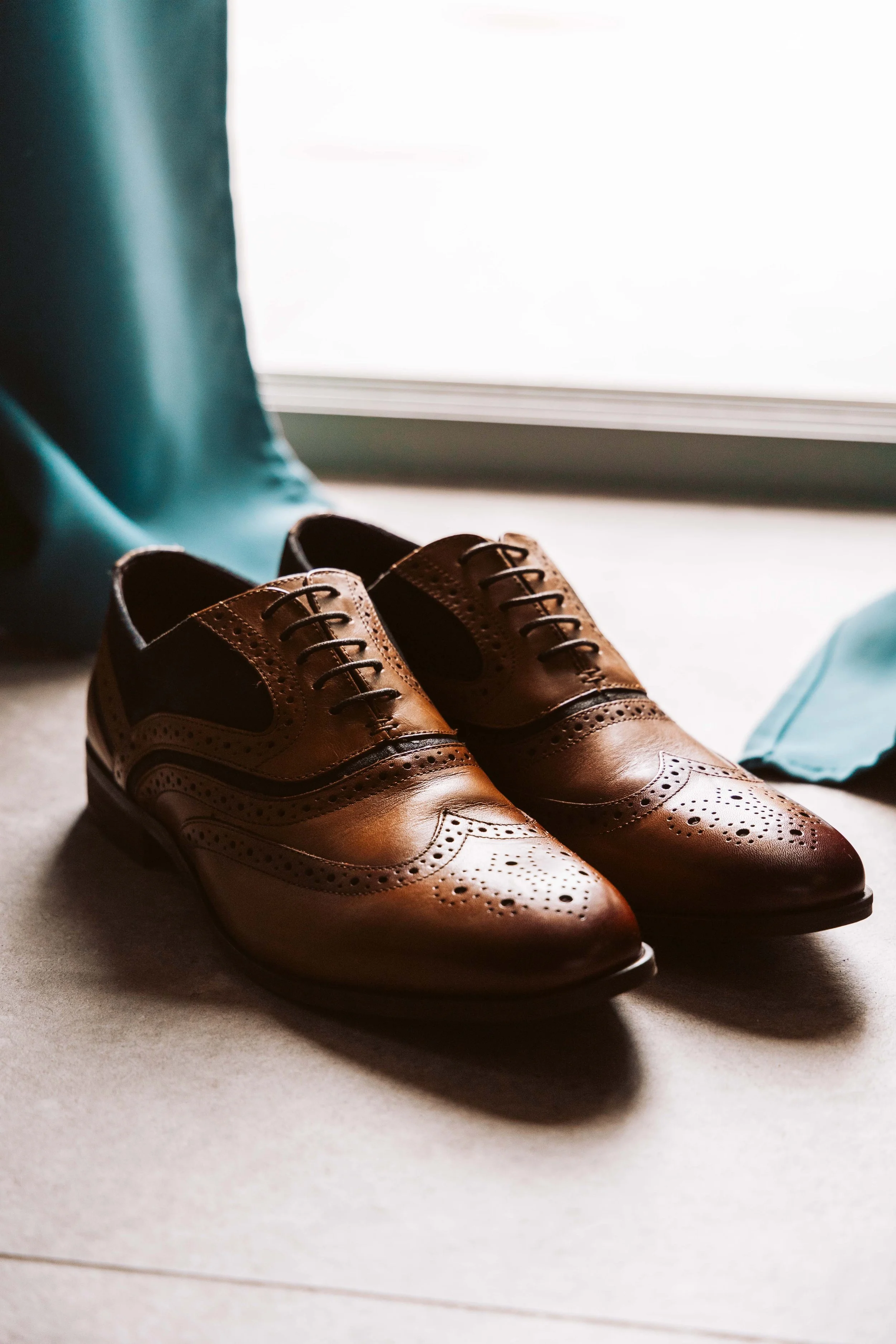 A pair of polished brown leather dress shoes placed on a light-colored floor near a window, with teal-colored curtains and a blue clothing item partially visible nearby.