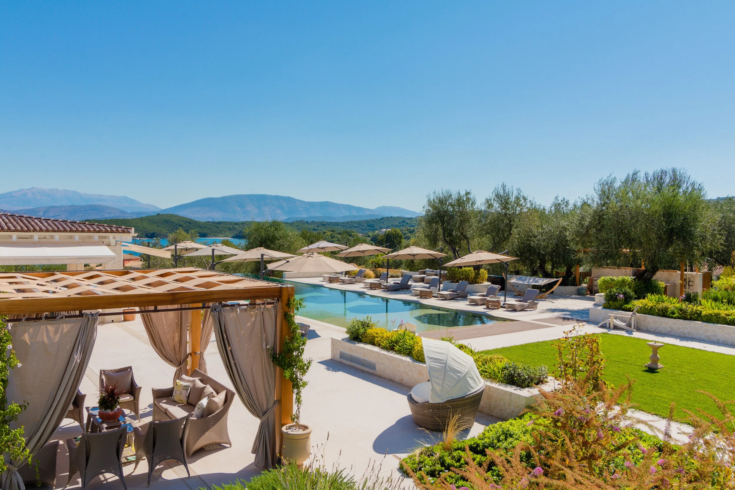 Luxury outdoor pool area with lounge chairs, umbrellas, and a shaded seating area on a sunny day, surrounded by greenery and mountains in the background.