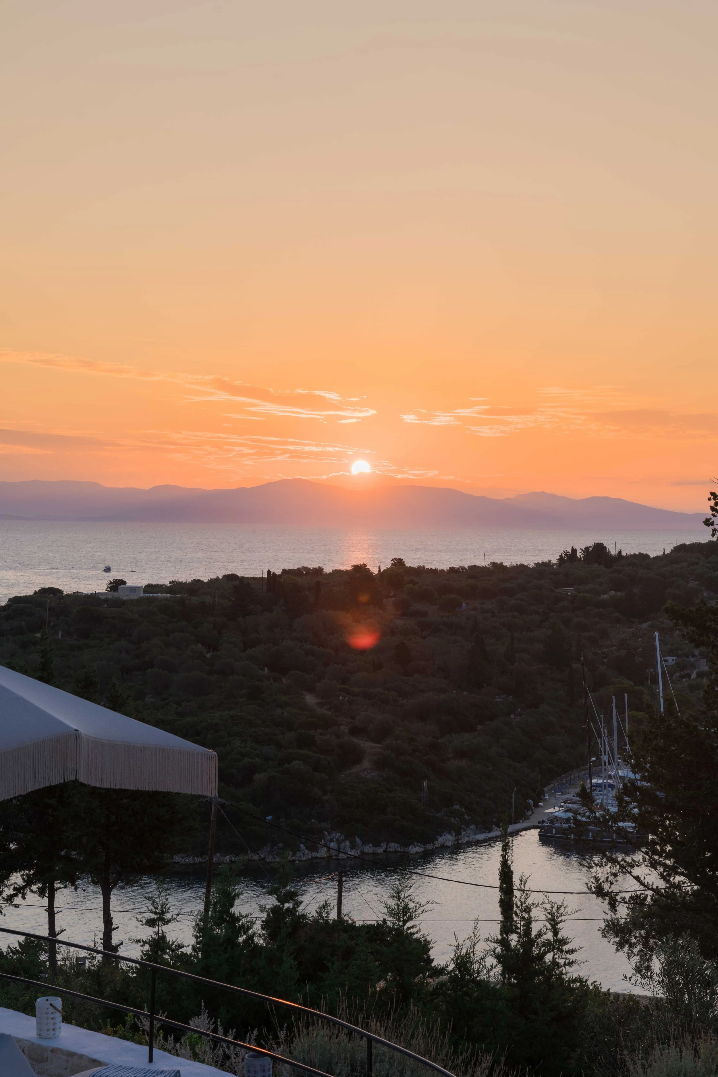 Sunset over a hilly coastal landscape with trees, boats anchored in a marina, and a body of water reflecting the sky's warm colors.