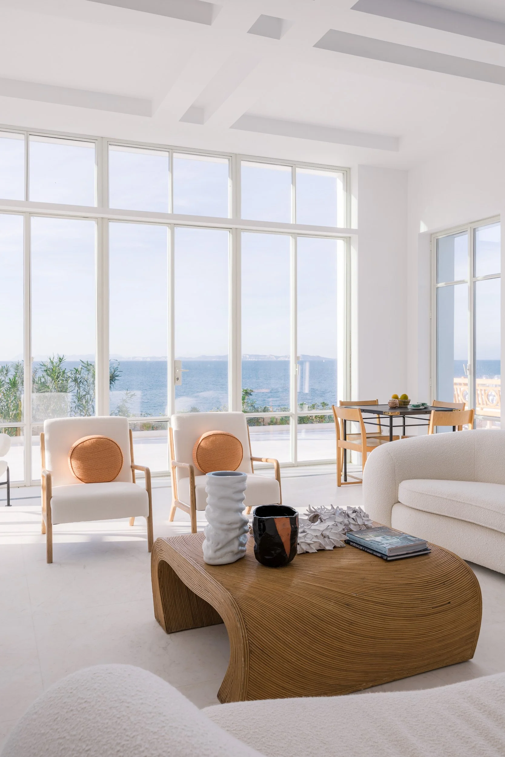 Modern living room with large windows overlooking the ocean, white walls, light-colored furniture, and decorative vases on a wooden coffee table.