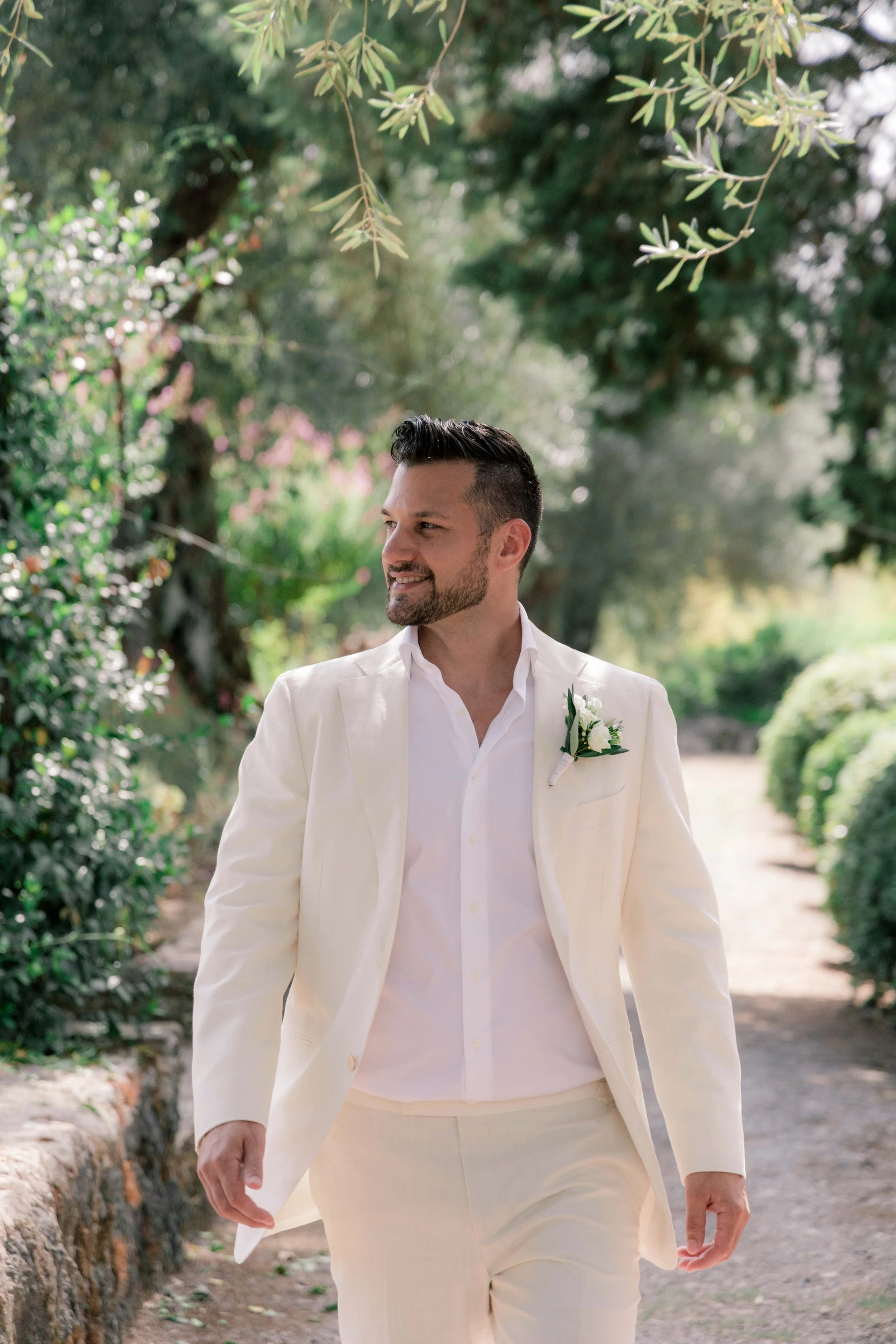 Man in white suit with boutonniere walking outdoors on a sunny day.