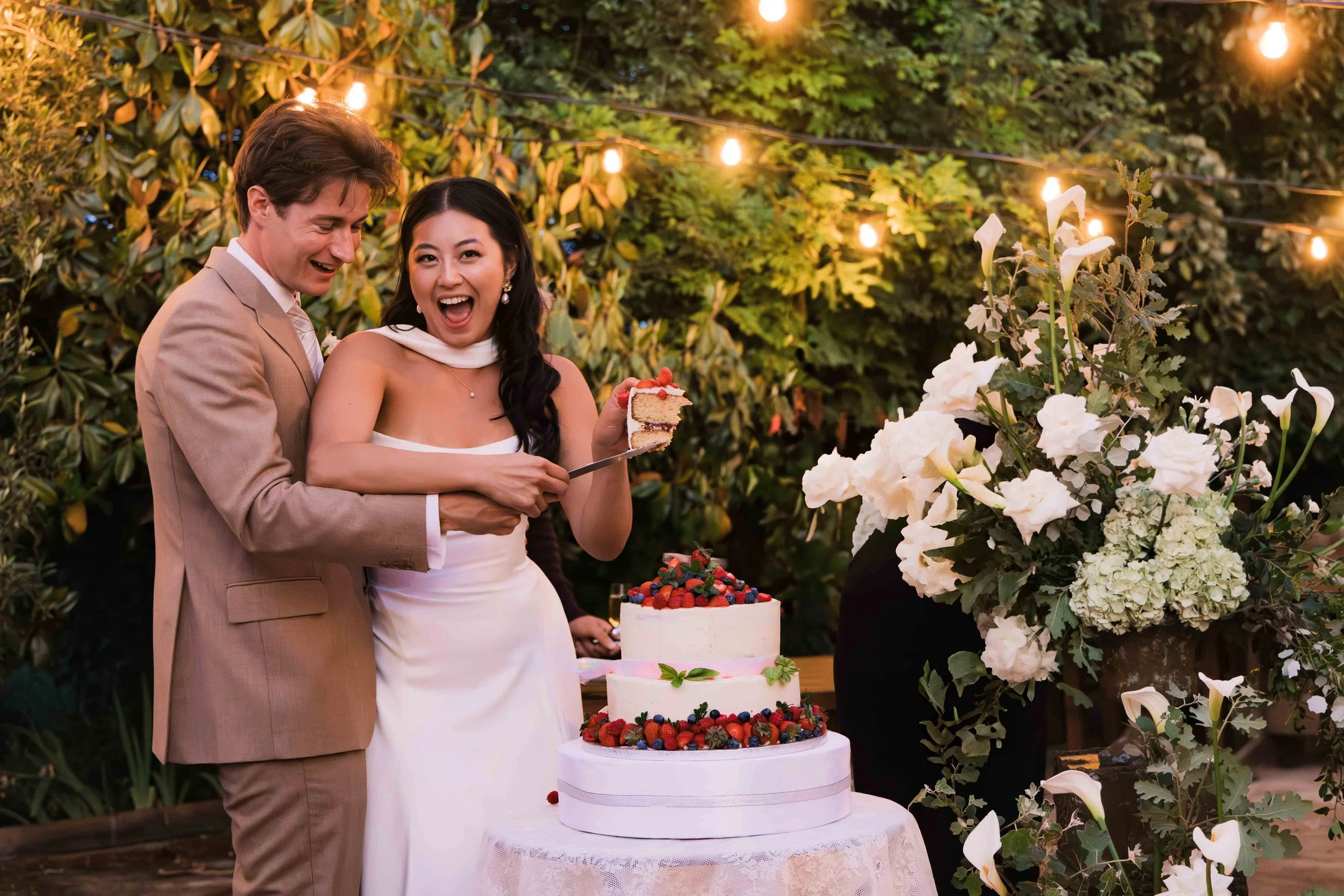 A newlywed couple cutting a wedding cake at an outdoor celebration, surrounded by floral arrangements and string lights.