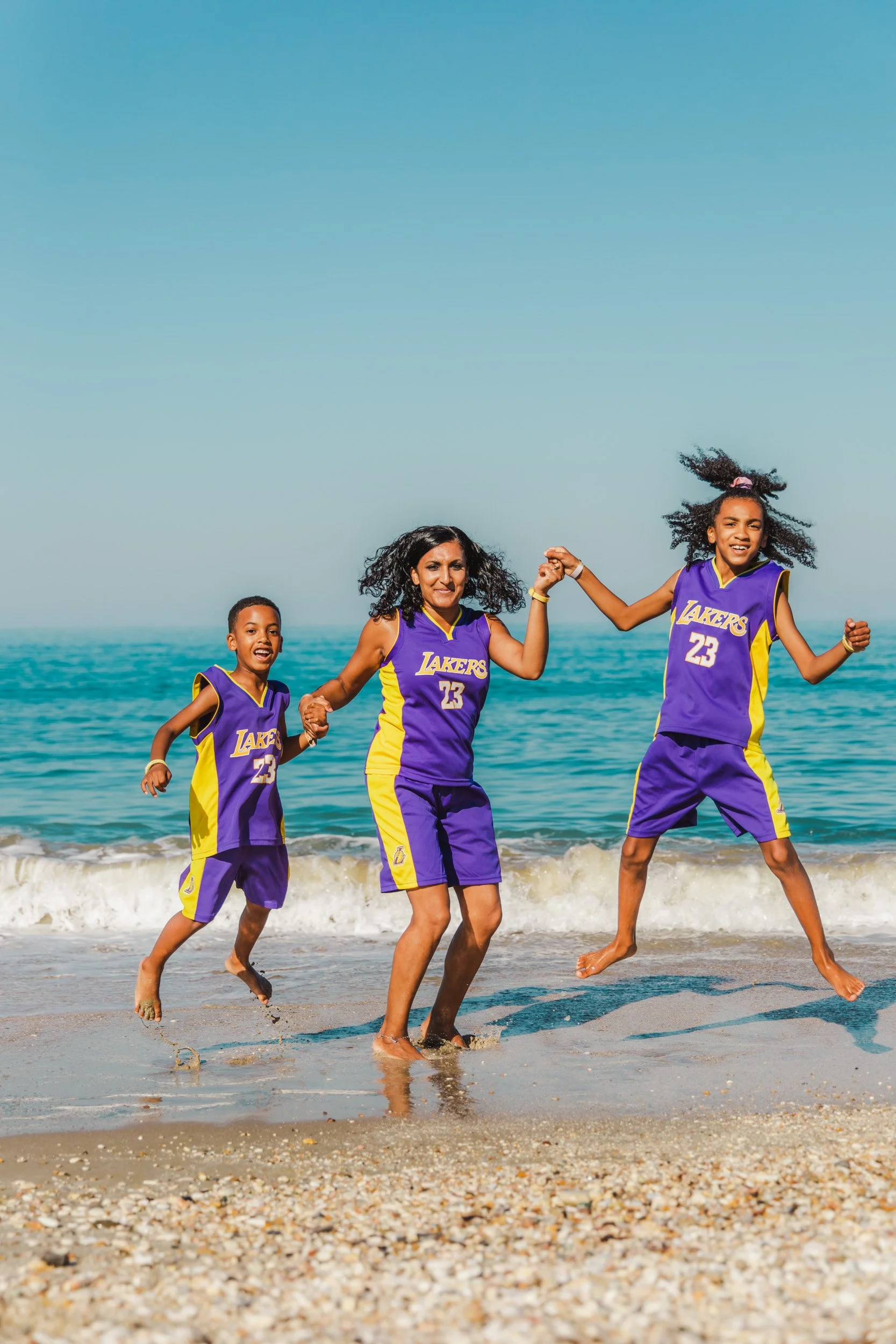 Three people in Los Angeles Lakers basketball uniforms playing and jumping on the beach with ocean waves in the background.