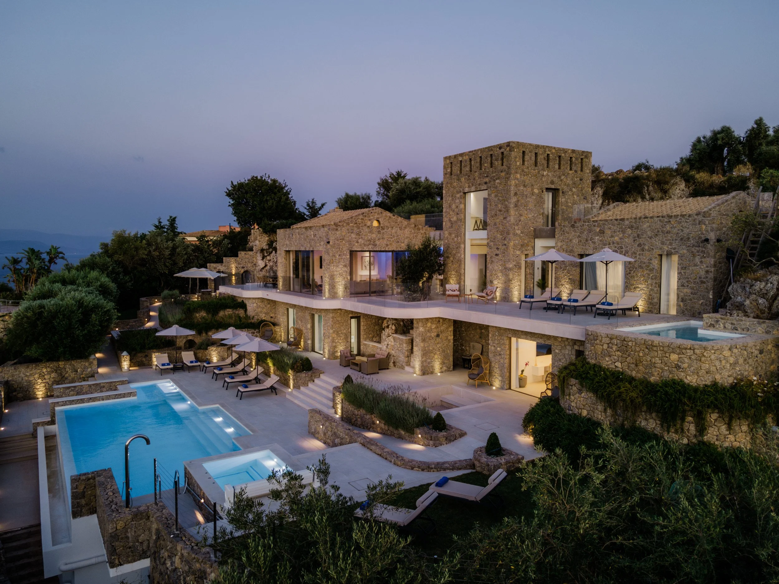Luxury stone villa with multiple levels and outdoor pools, surrounded by greenery, at dusk.