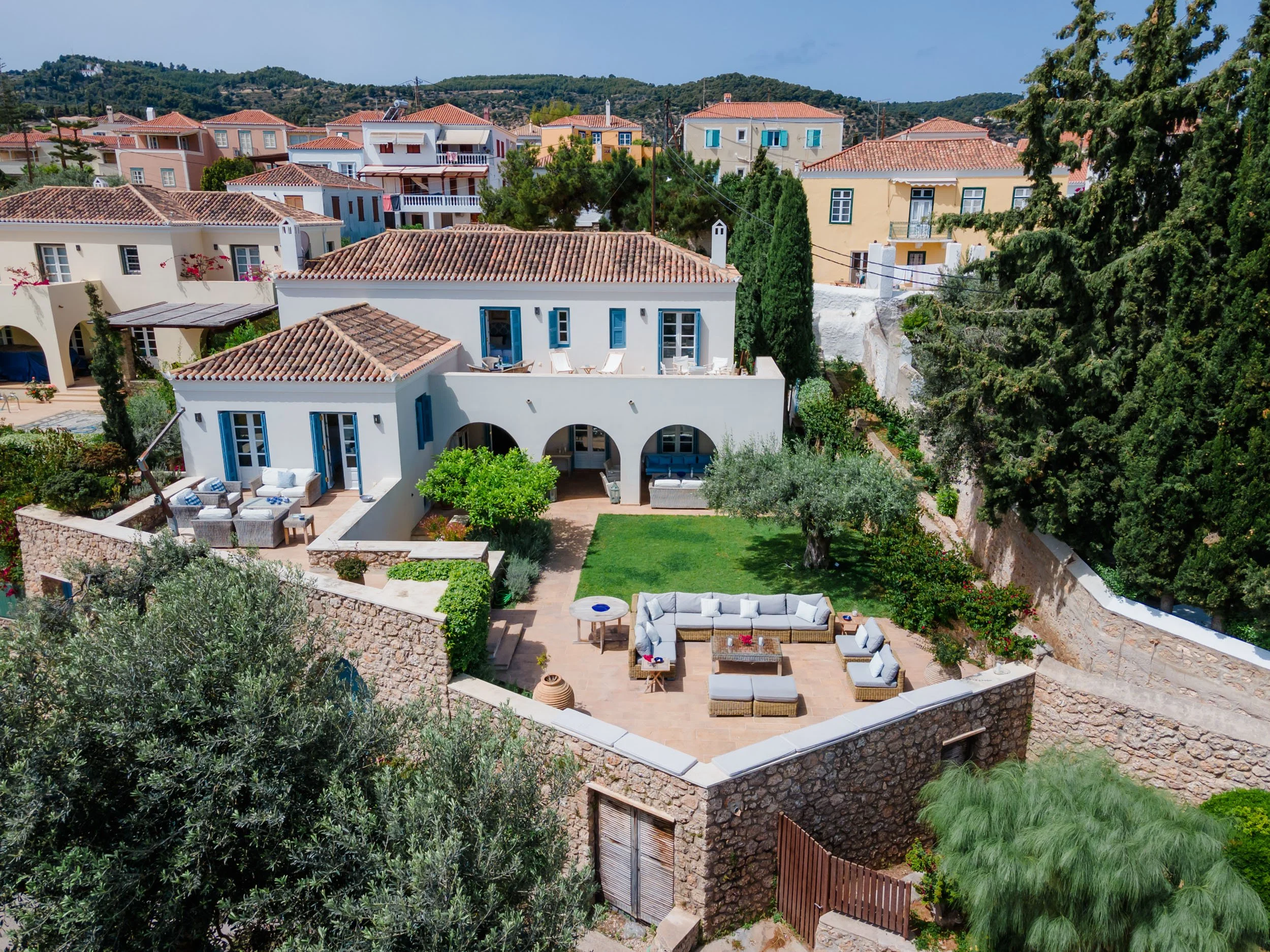 A white house with a red tile roof and blue shutters, featuring outdoor seating areas and a garden, in a neighborhood with similar style houses surrounded by greenery.