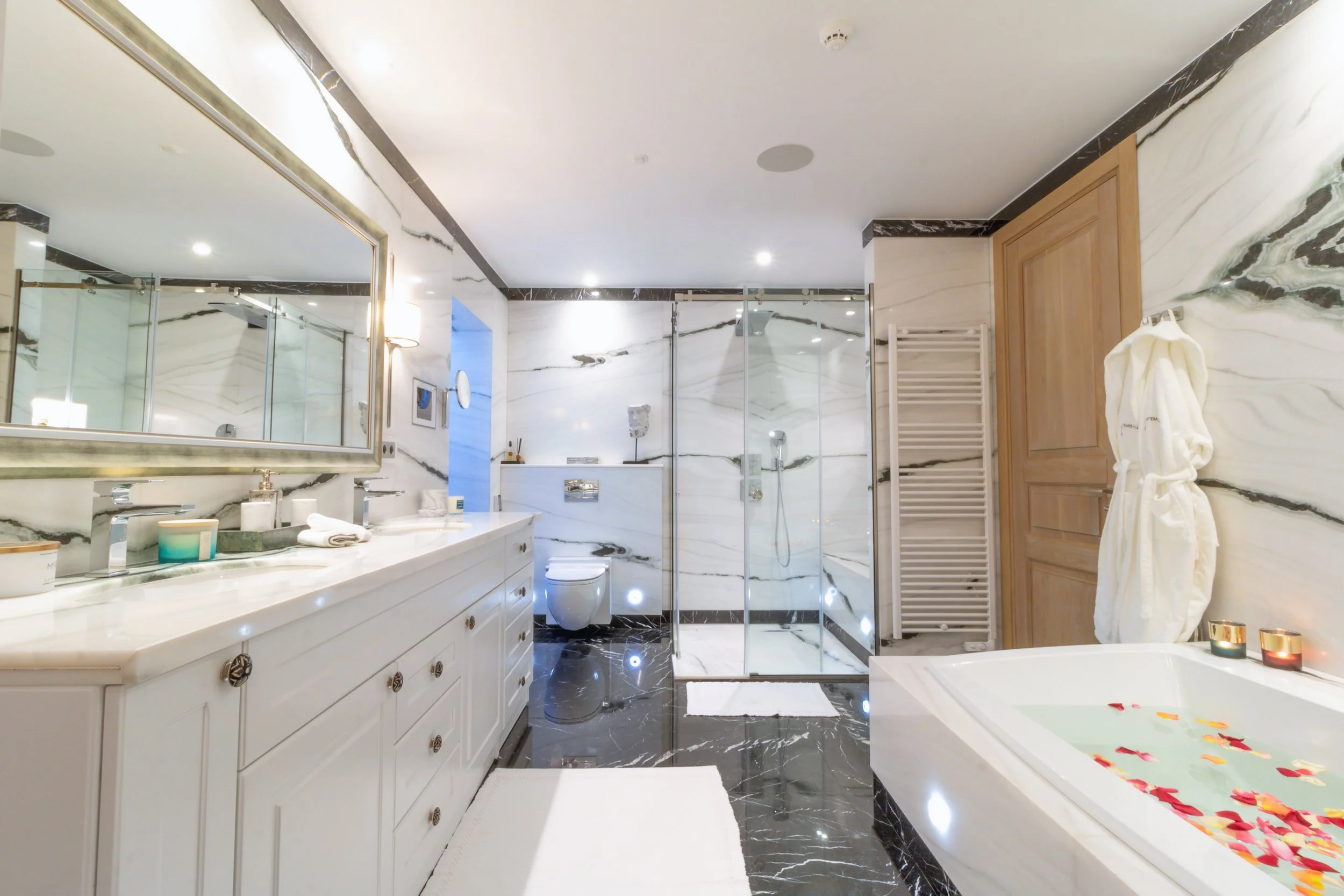 Luxury bathroom with marble walls and floor, double vanity with large mirror, walk-in shower with glass doors, bathtub filled with water and flower petals, and a wooden cabinet.