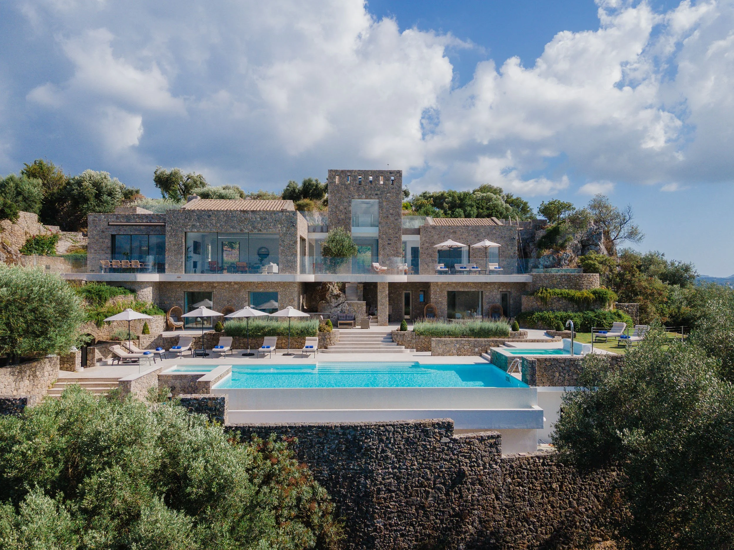 Luxury modern house built into a hillside with a large swimming pool and outdoor lounge areas under umbrellas, surrounded by greenery under a partly cloudy sky.